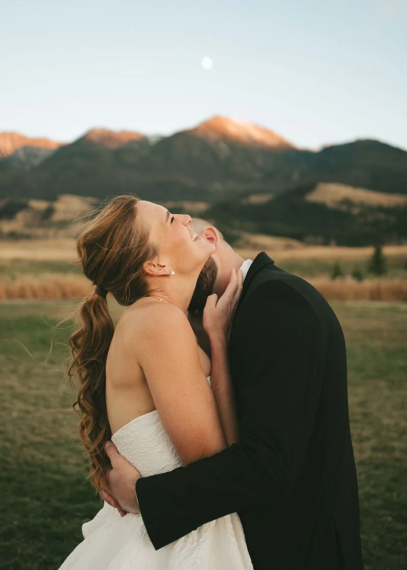 A bride and groom embrace outdoors at sunset with Montana mountains in the background, the bride smiling and leaning her head back.