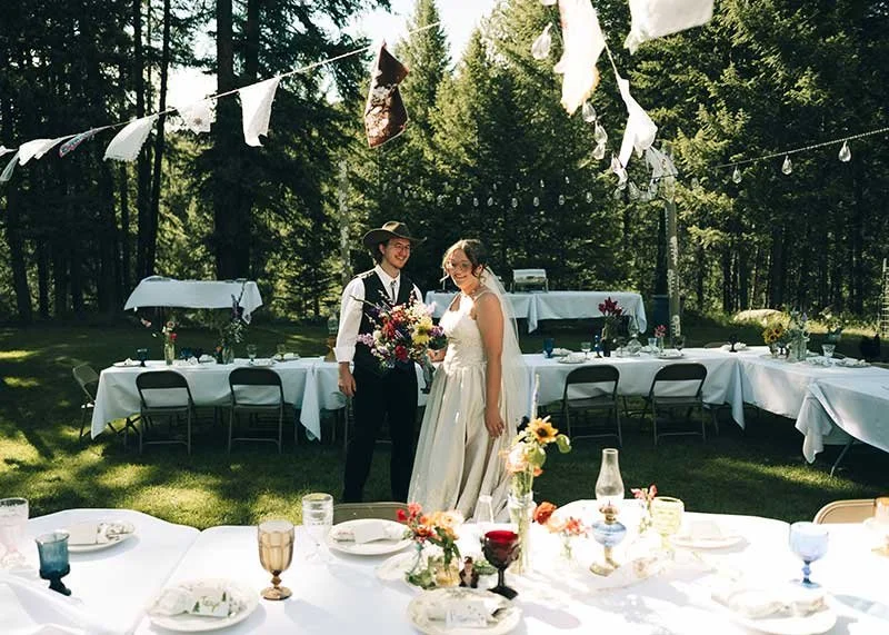A Montana bride and groom standing outdoors under hanging decorations, smiling, with a decorated table in front of them, greenery, and trees in the background.