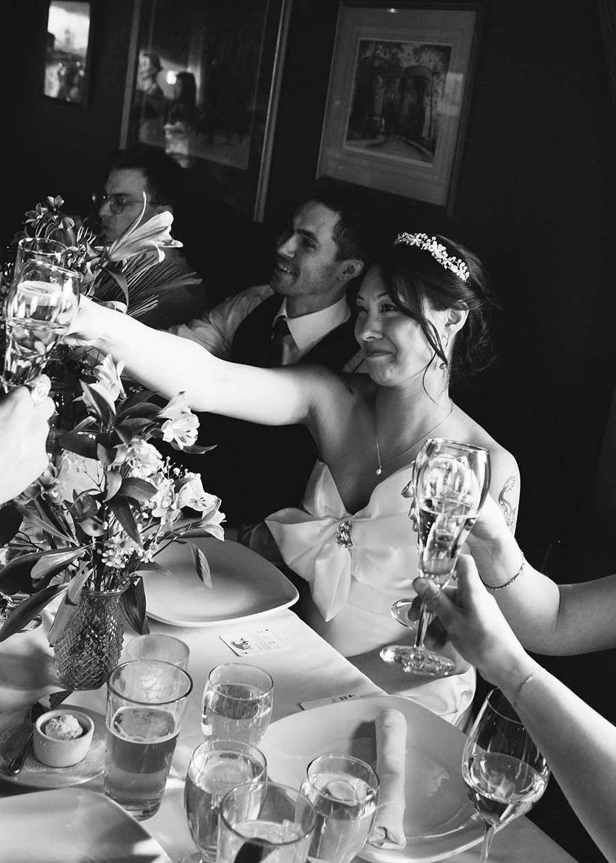 A bride and groom at their Montana wedding reception raising glasses for a toast, surrounded by friends and family, with floral arrangements and dinner settings on the table.