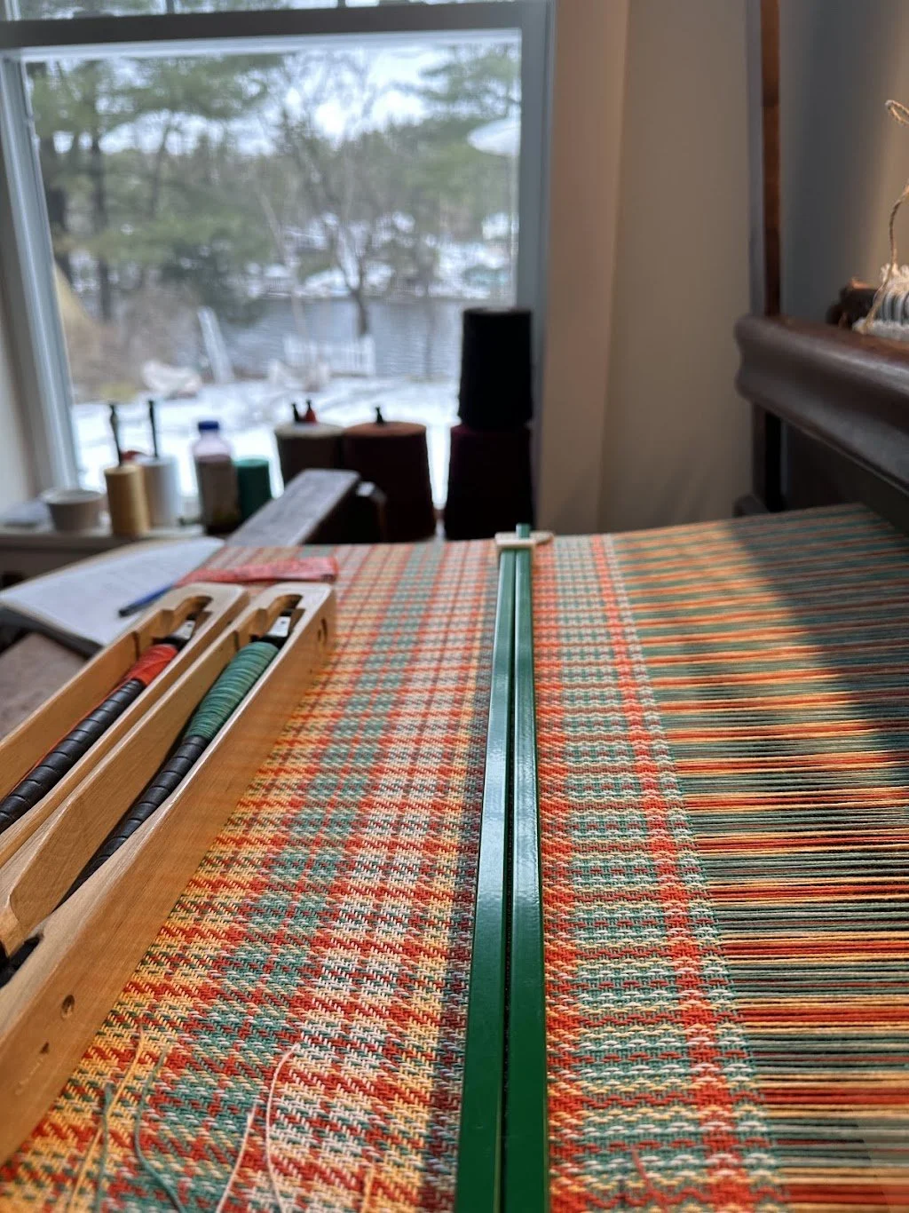 Interior view of a room with a large window overlooking a wintery outdoor scene, featuring trees and snow. On a table, there is a long green ruler, spools of thread, and sewing supplies, with a fabric that has a colorful pattern.