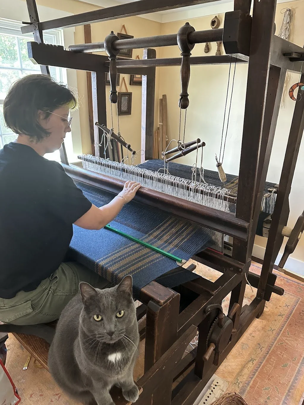 A woman is working on a large traditional loom in a cozy room with natural light, while a gray cat sits on the table nearby, looking at the camera.