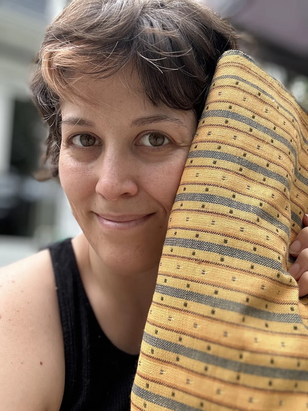 Close-up of a woman with short brown hair and hazel eyes holding a yellow and gray striped pillow, smiling gently indoors.