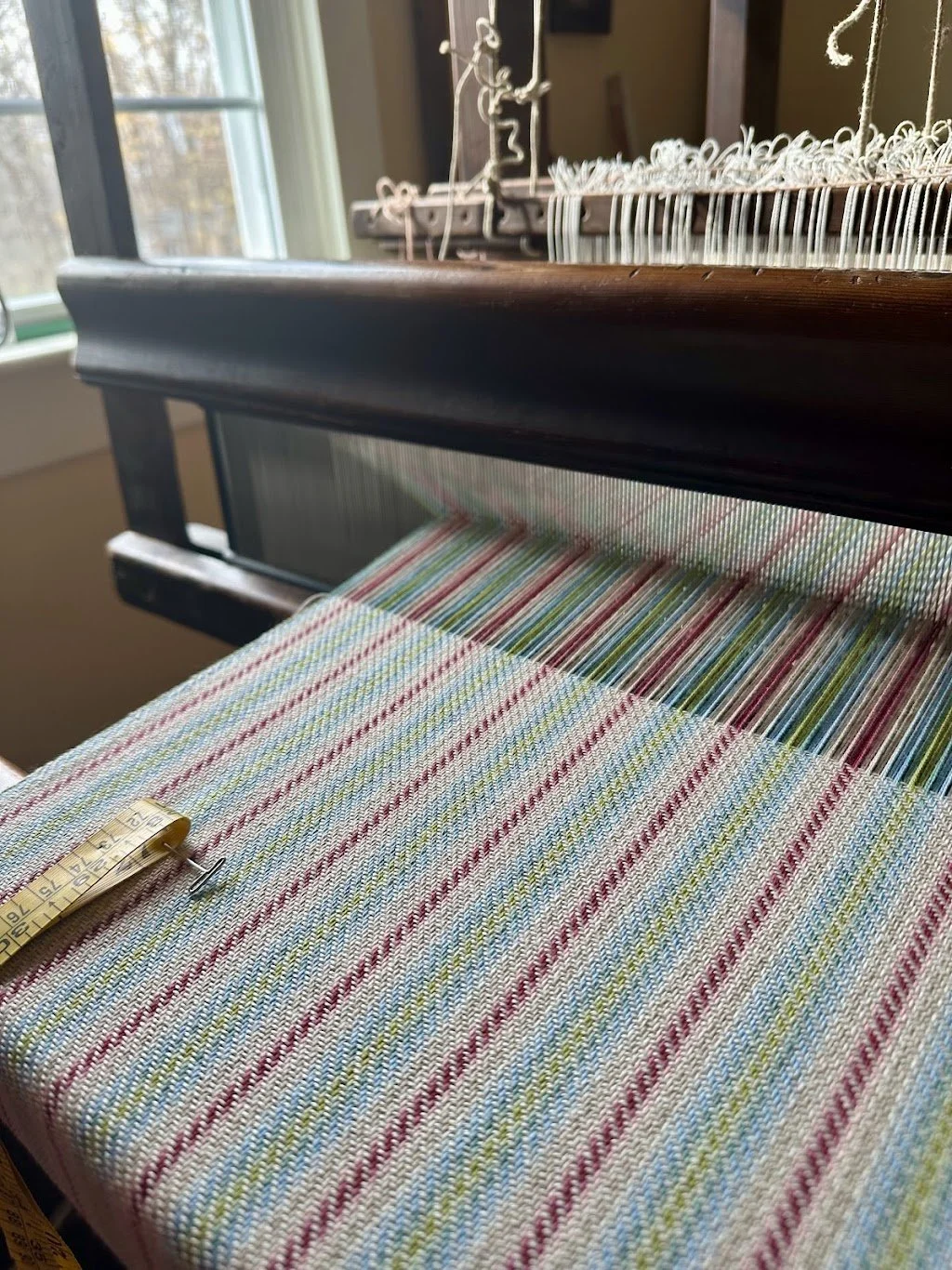 A colorful striped woven fabric being woven on a loom with a measuring tape placed on it, in a room with a window.