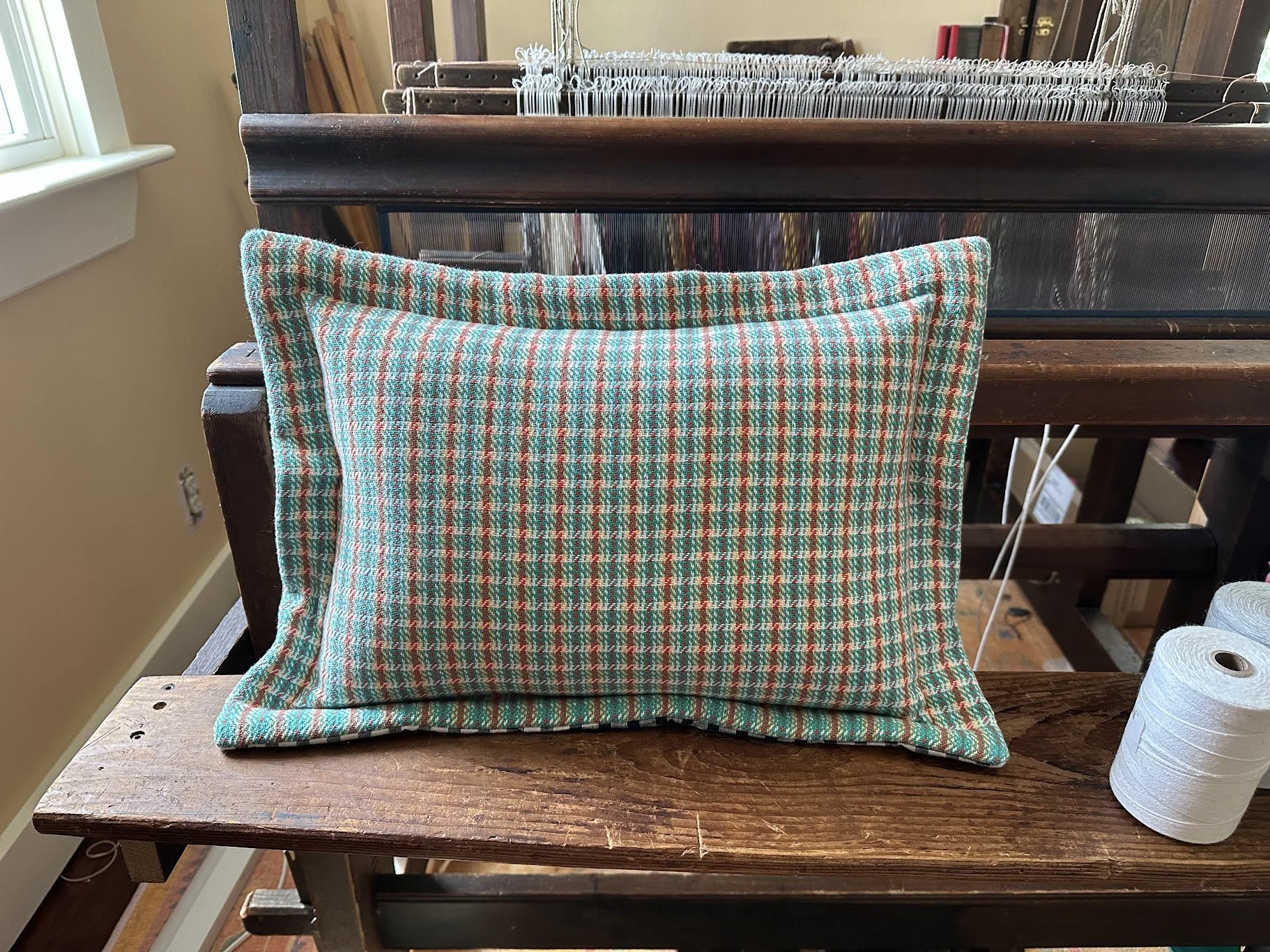 A rectangular pillow with a multicolored plaid pattern resting on a wooden surface in front of a vintage loom. A roll of white thread sits on the right side of the pillow, next to the loom.
