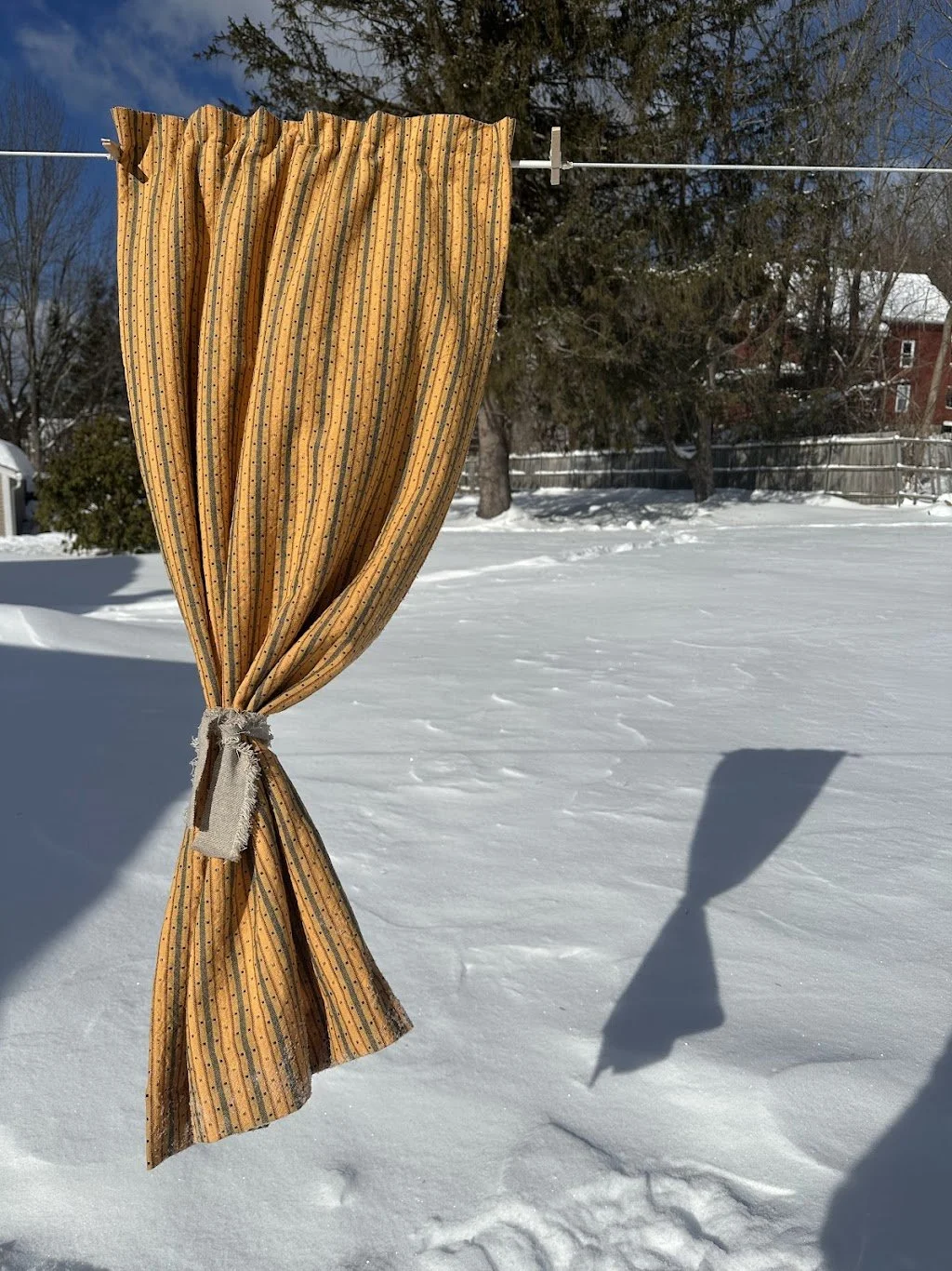 A brown striped curtain tied back and hanging on a clothesline in a snowy backyard with trees and buildings in the background.