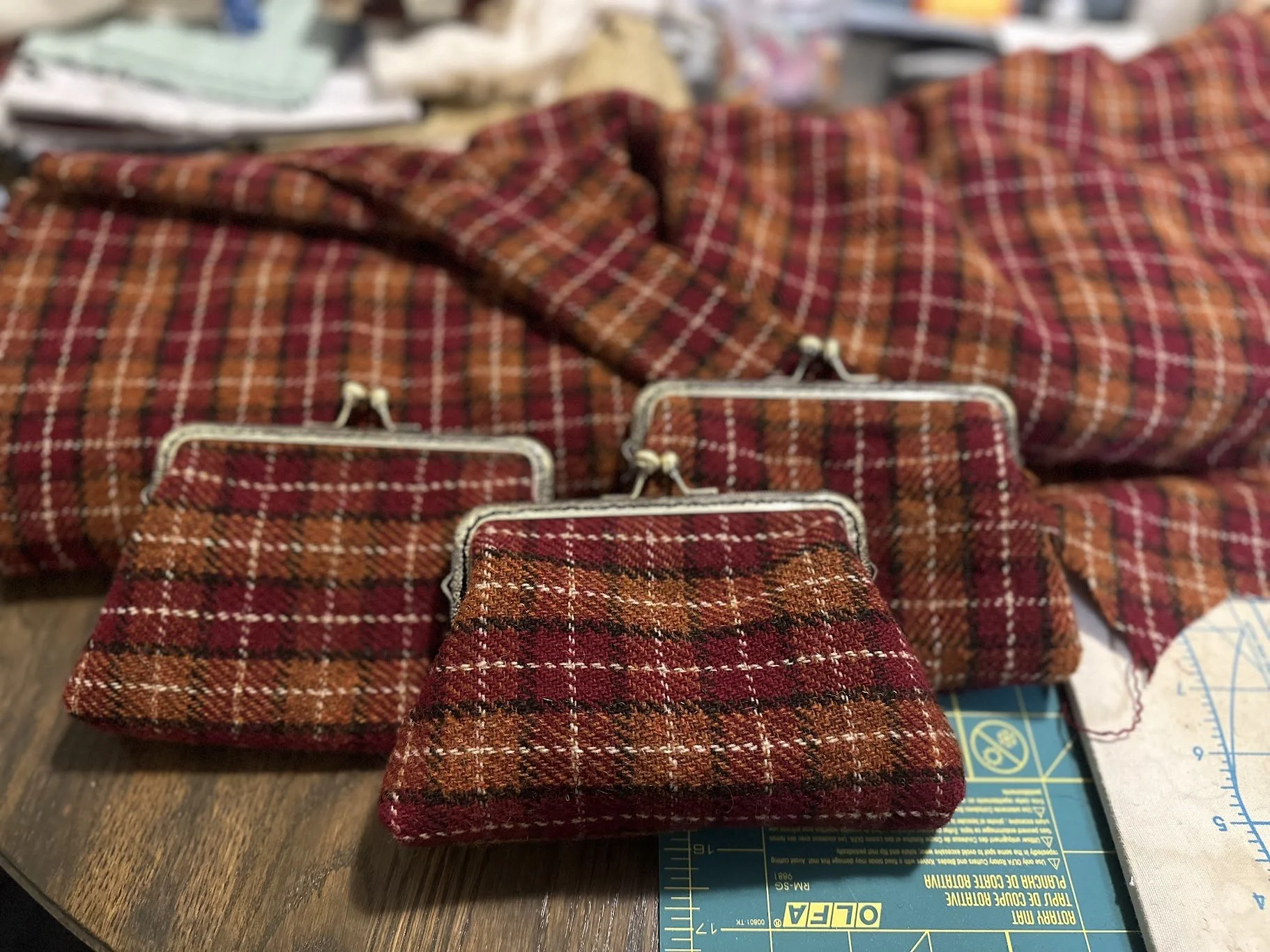 Multiple small fabric pouches with metal clasps featuring red and orange tartan pattern, placed on a wooden surface with a cutting mat and fabric in the background.