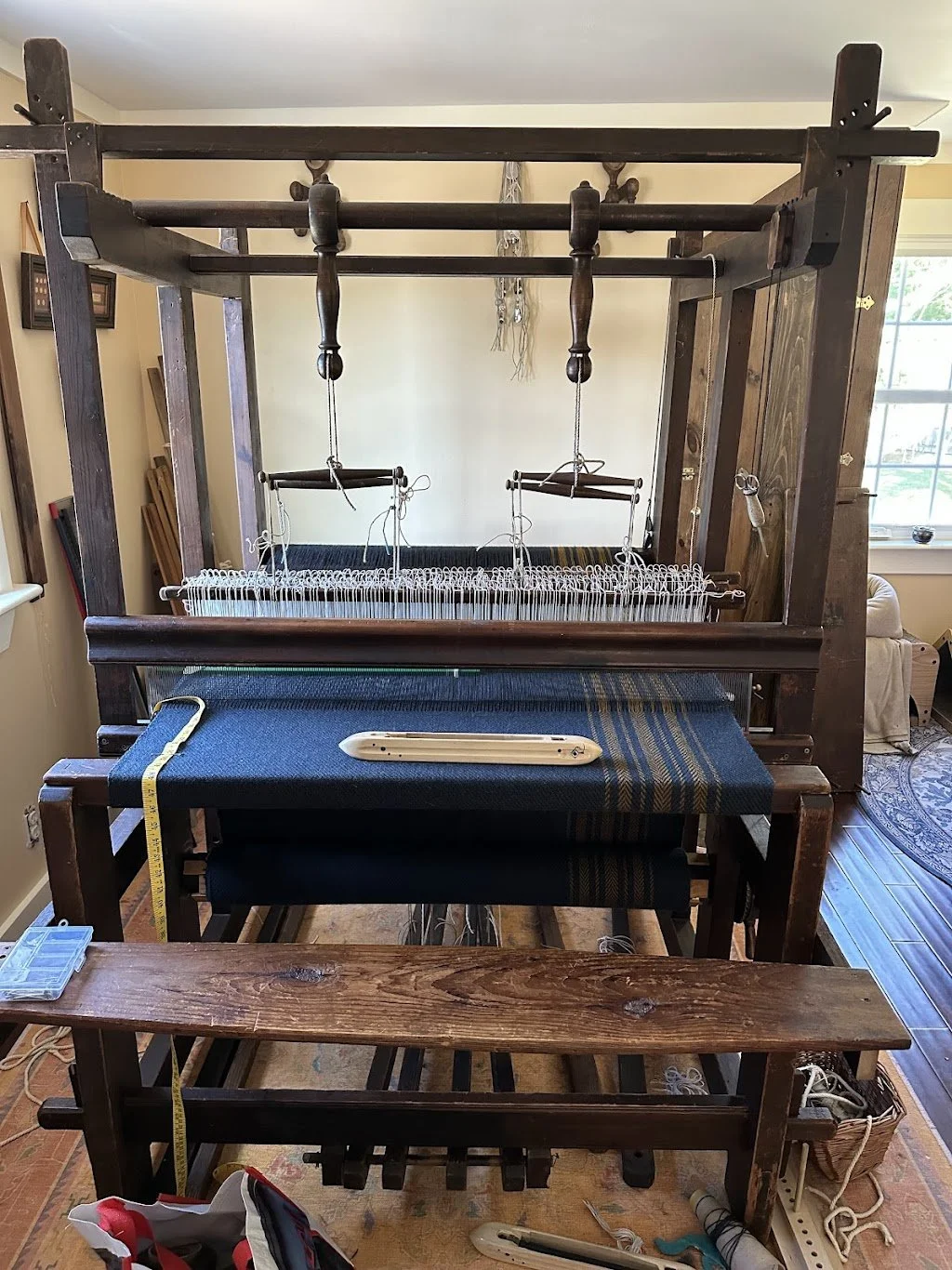 A large, vintage wooden loom in a room with natural light from a window, with tools and measuring tape nearby.