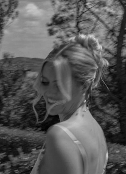 A black and white photo of a woman with blonde hair styled in an updo, smiling with her eyes closed outdoors, with trees and a cloudy sky in the background.