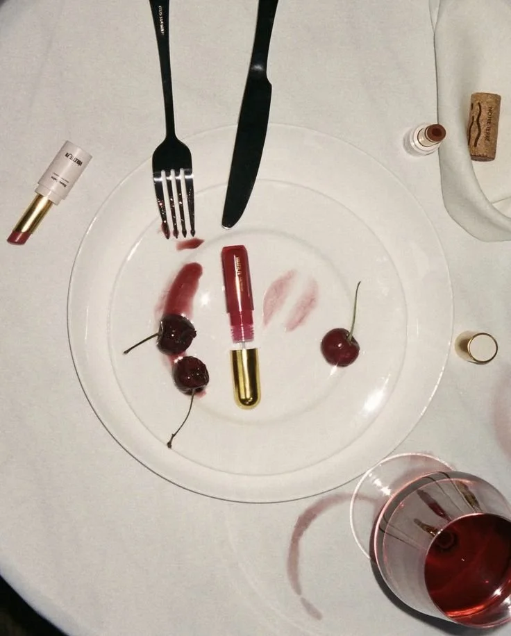 Empty transparent plate with a lipstick mark, cherries, and lipstick tubes. A glass of red wine is nearby on a white tablecloth, with lipstick and nail polish bottles and makeup tools around.