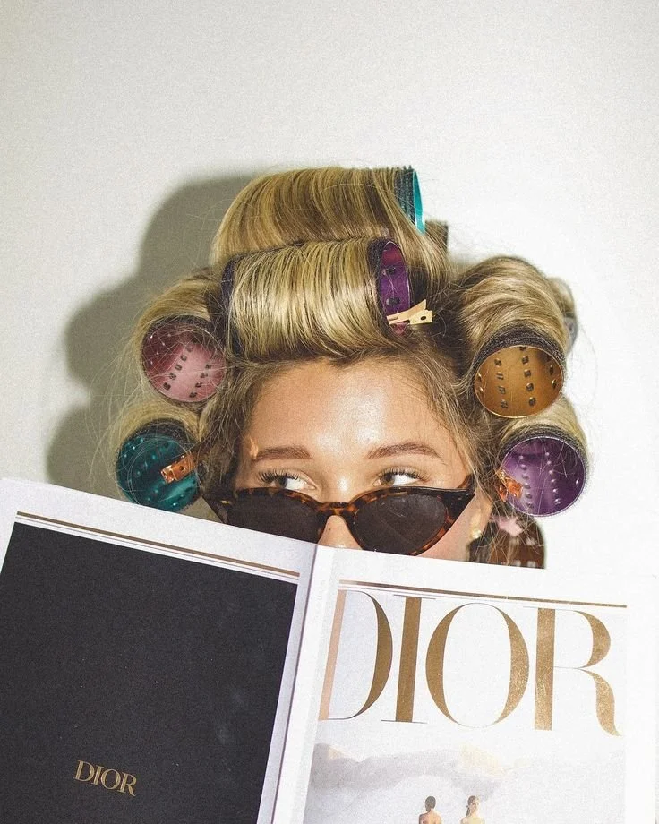 A woman with blonde hair in rollers, wearing sunglasses, reading a Dior magazine.