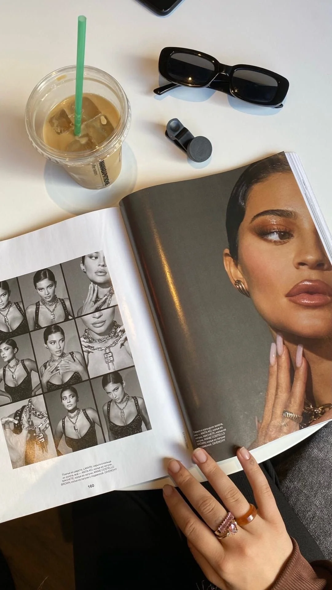 A flat lay of a magazine open to a fashion photo and black-and-white photos of a woman wearing jewelry, a pair of black sunglasses, a small black device, and a plastic cup with iced coffee and a green straw on a white table.
