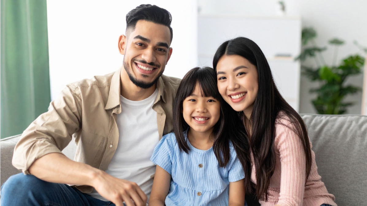 Une famille représentant un père, une mère et une fille souriants assis sur un canapé dans un salon lumineux.