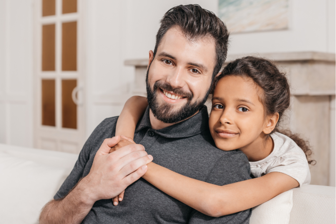 Un homme et une jeune fille souriante, assis sur un canapé, en train de se faire un câlin, dans une ambiance chaleureuse et familiale.