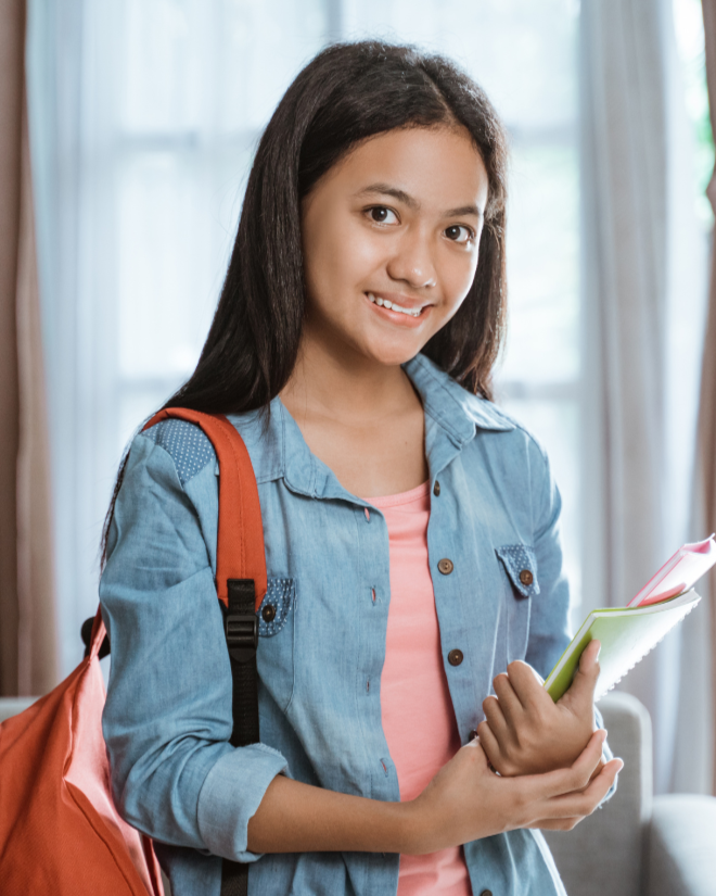 Jeune fille souriante avec un sac à dos orange, tenant des cahiers colorés, dans une pièce avec des fenêtres lumineuses.