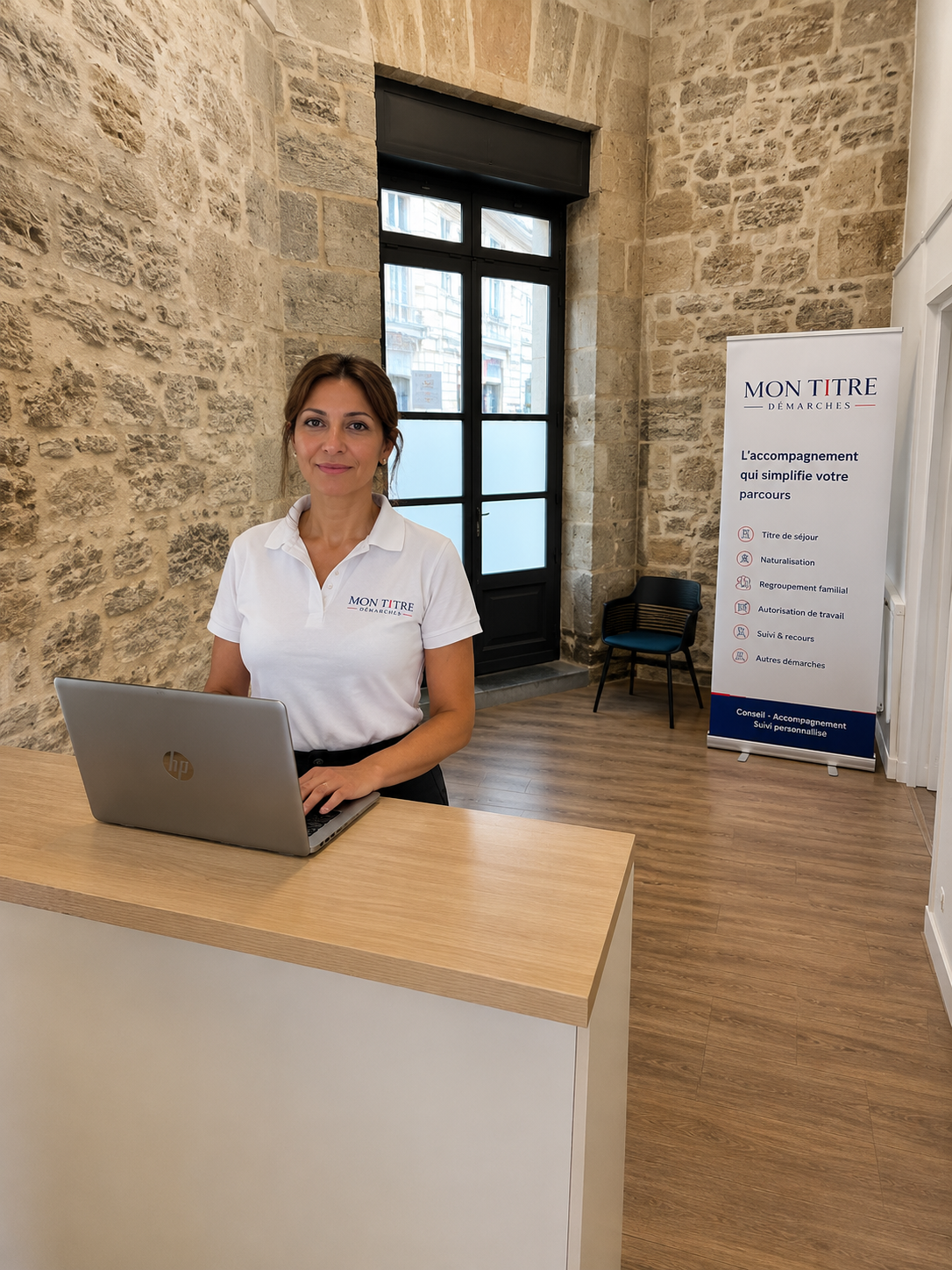 Une femme souriante portant un polo blanc avec le logo « Mon Titre Démarches » assise derrière un bureau avec un ordinateur portable HP. Il y a une affiche informative à droite mentionnant différents services offerts, dans une pièce avec murs en pierre et un sol en bois.