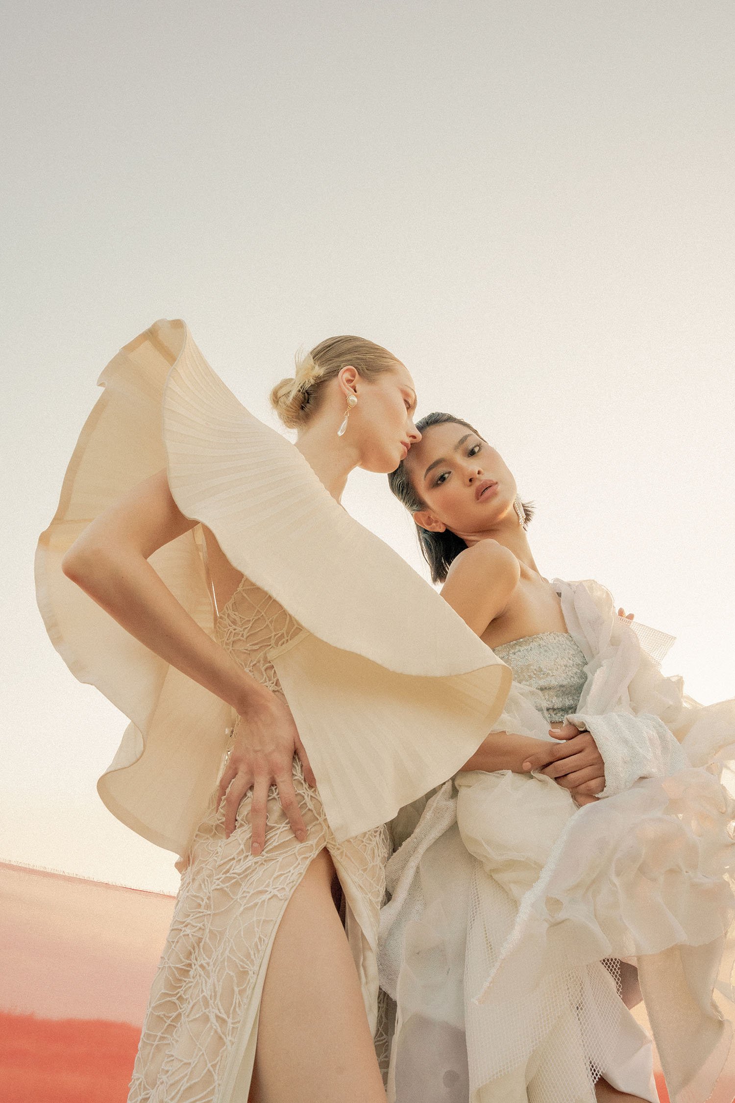 Two women dressed in fashionable, beige and white outfits, posing outdoors against a pale sky.