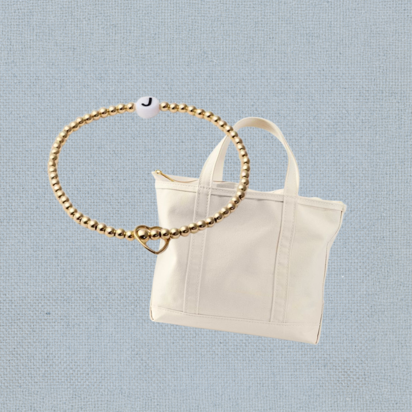 A white tote bag with handles and a gold zipper, with a gold beaded bracelet featuring a heart-shaped charm and a bead with the letter 'J' floating above it, against a light blue textured background.