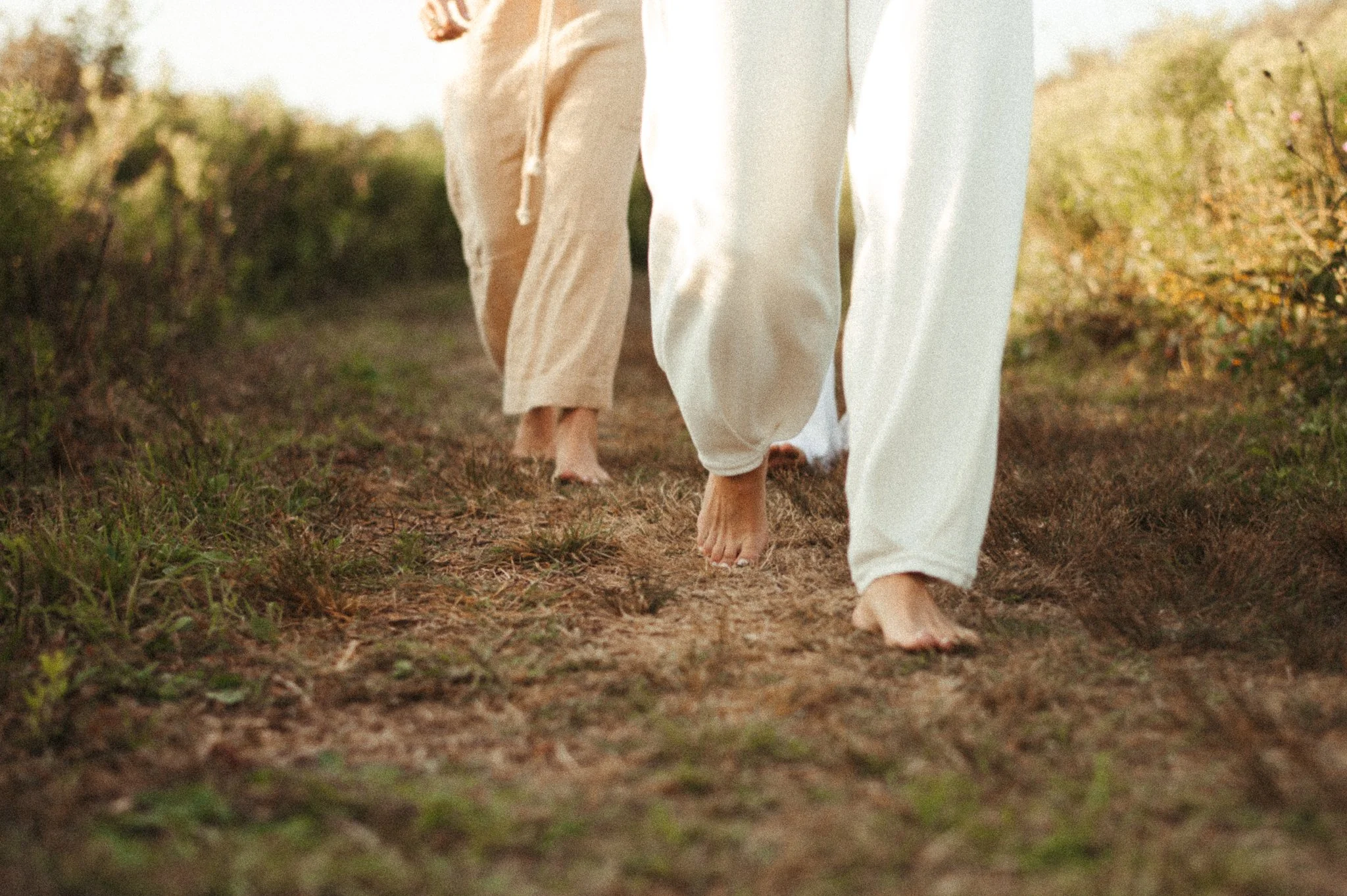 Three people walk barefoot along a dirt trail through a grassy, bushy area during daylight. They wear loose, comfortable clothing in neutral colors, with only their lower bodies and feet visible.