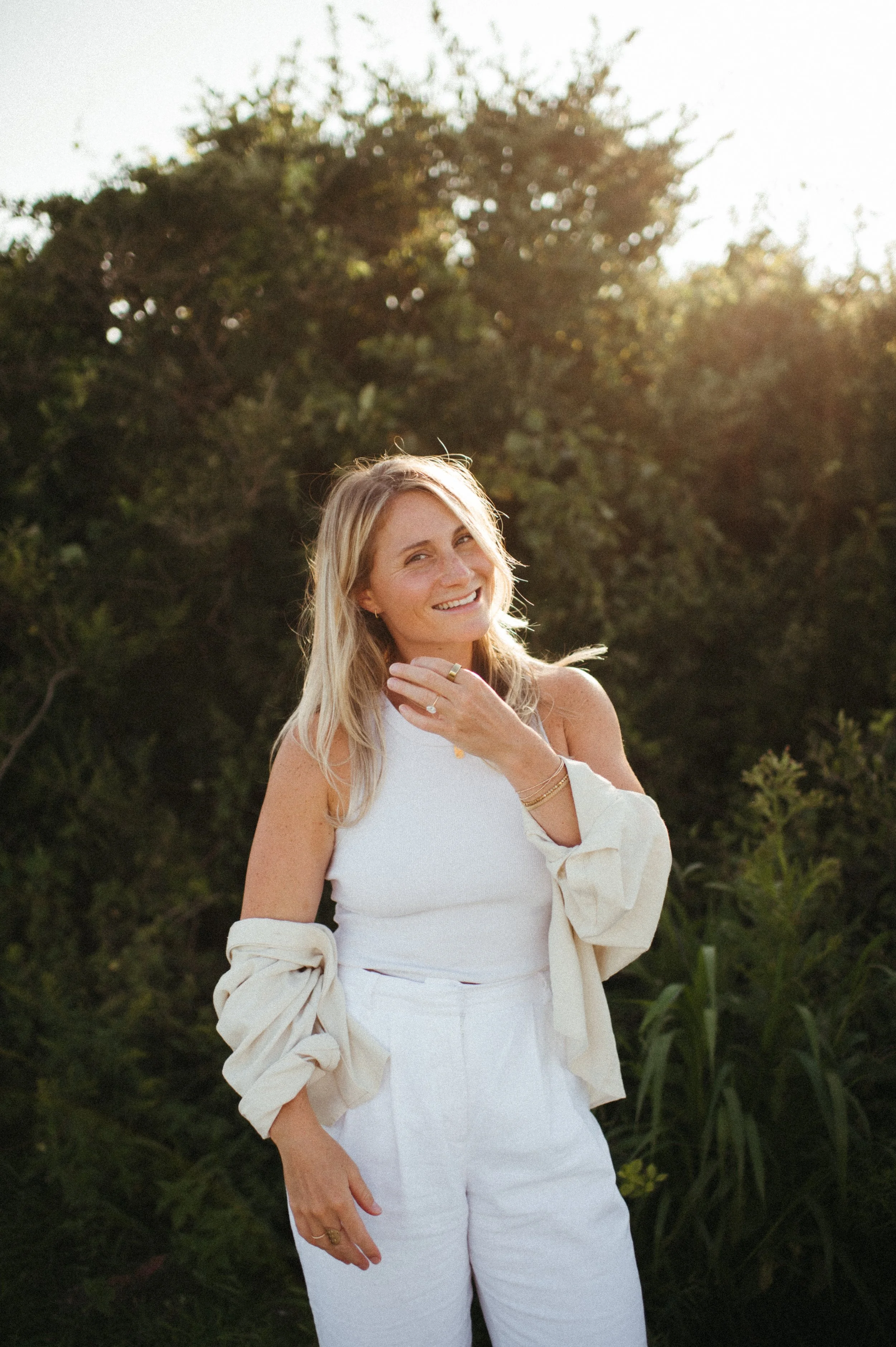 A woman with blonde hair smiling outdoors, wearing a white sleeveless top, white pants, and a light-colored jacket on her arms, with greenery and sunlight in the background.