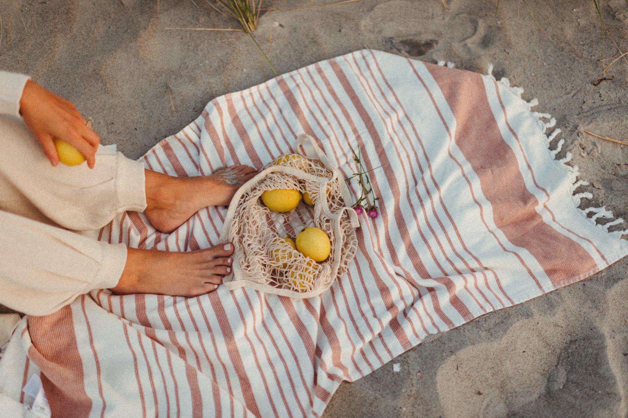 View from above of a person with bare feet sitting on a wide striped beach towel on the sand with a small net bag containing three yellow eggs and a small pink flower nearby.