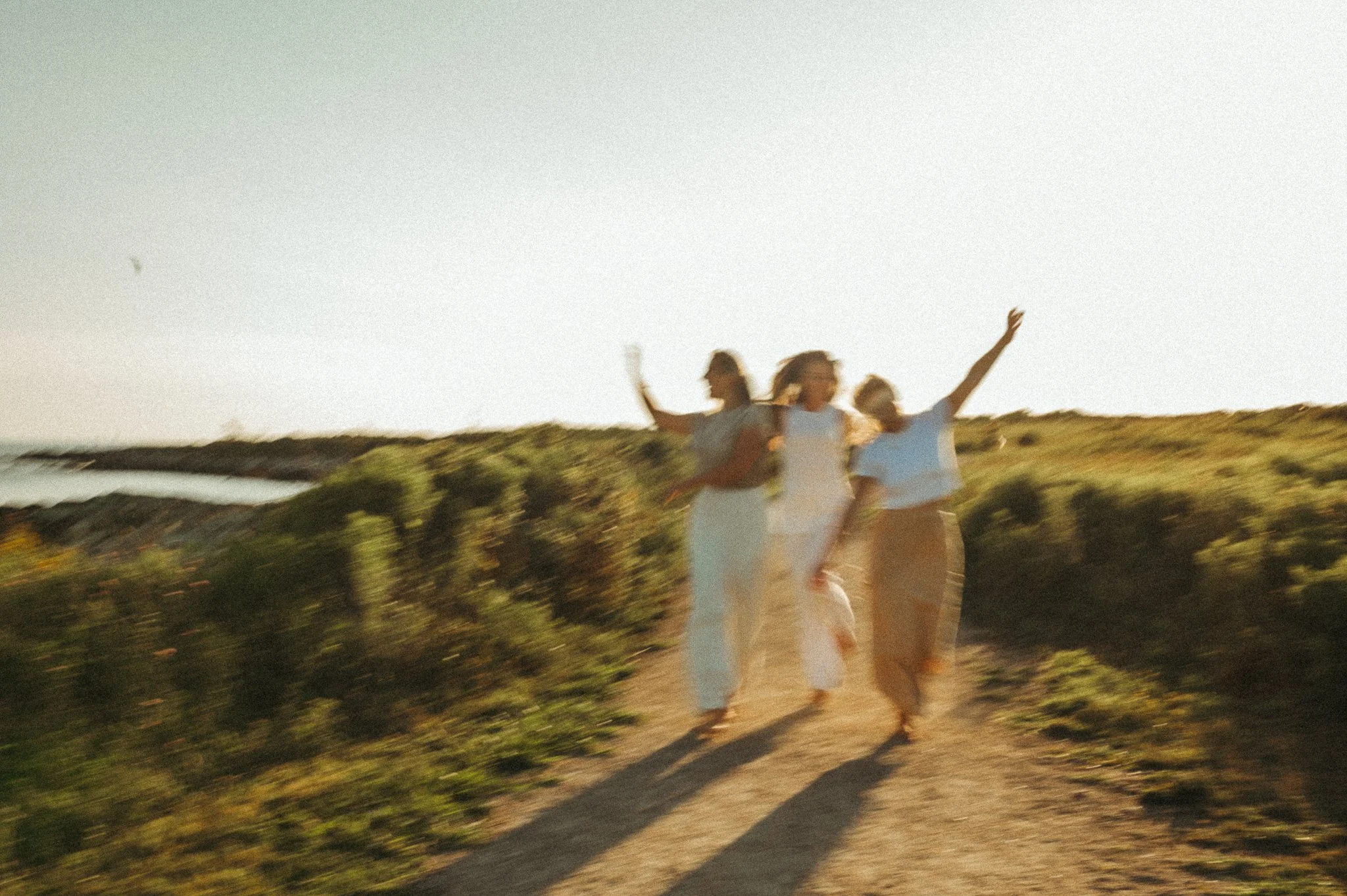 Three women walking on a dirt path along a coastal landscape, enjoying a sunny day.