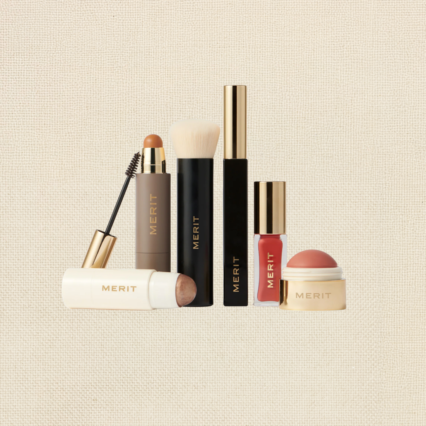Collection of Merit beauty products including a brow gel, foundation stick, makeup brush, lip gloss, and lip balm arranged on a neutral textured background.