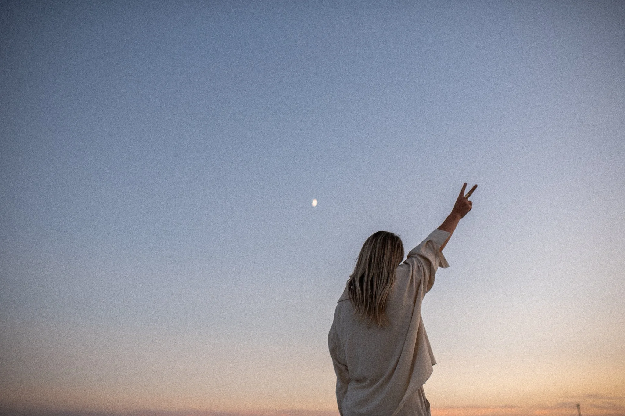 A woman with long hair making a peace sign with her hand, facing away, in a sunset or sunrise sky with a visible moon.