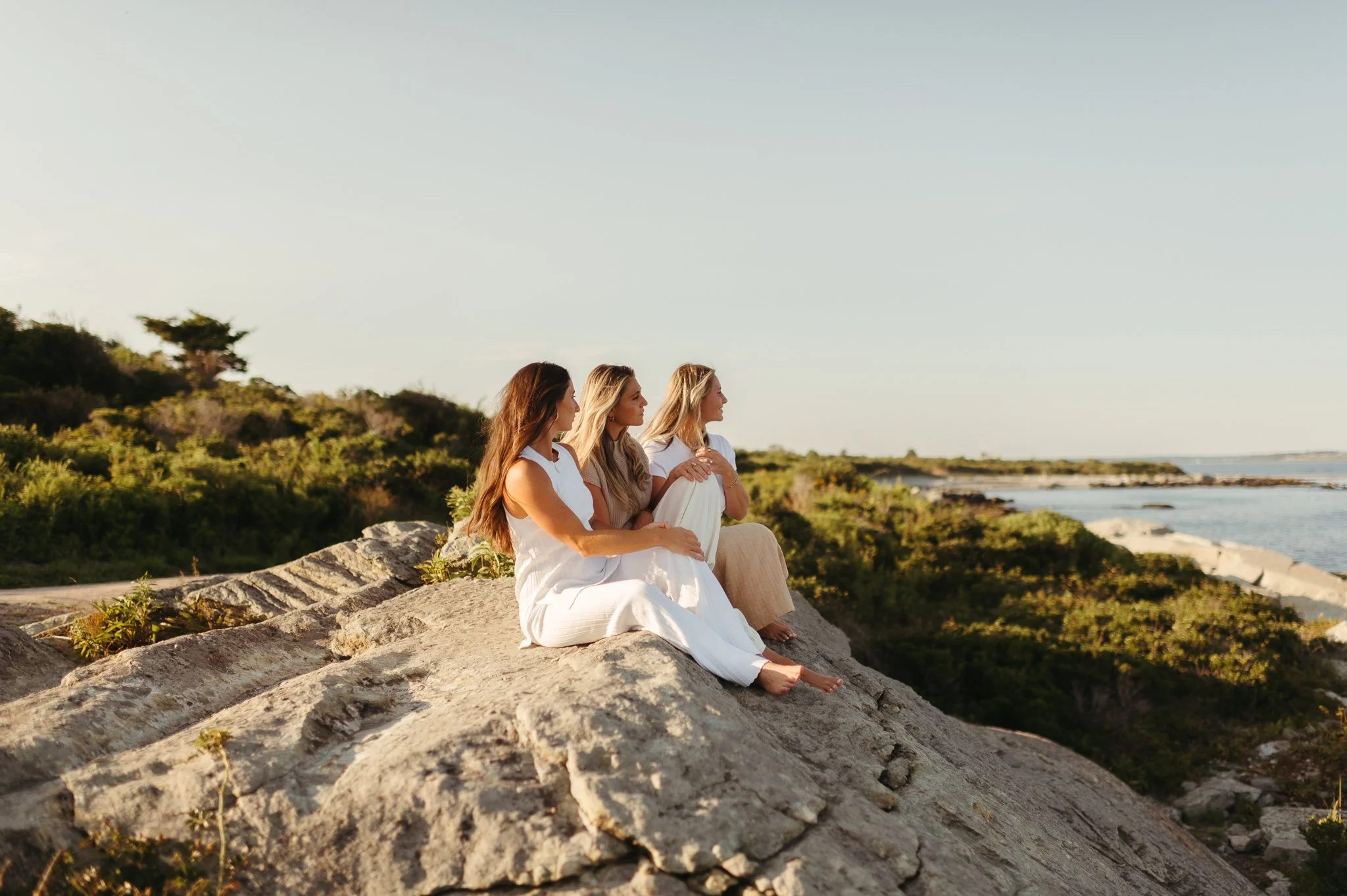 Three women sitting on a large rock on the coast, gazing at the ocean during sunset.