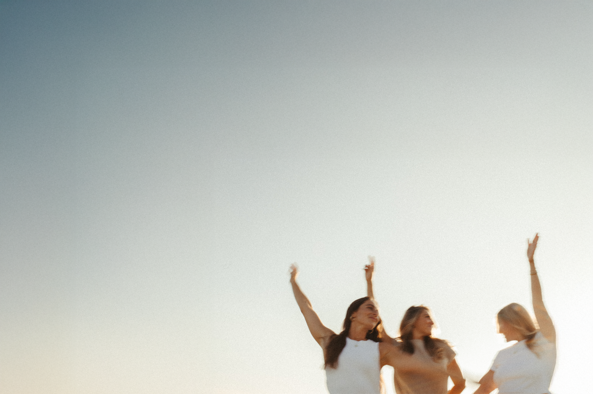 Three women with long hair and light skin outside at sunset, celebrating with raised arms.
