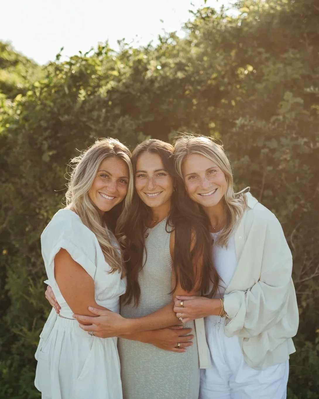 Three women standing outdoors in front of green foliage, smiling and embracing each other, with sunlight shining from behind.
