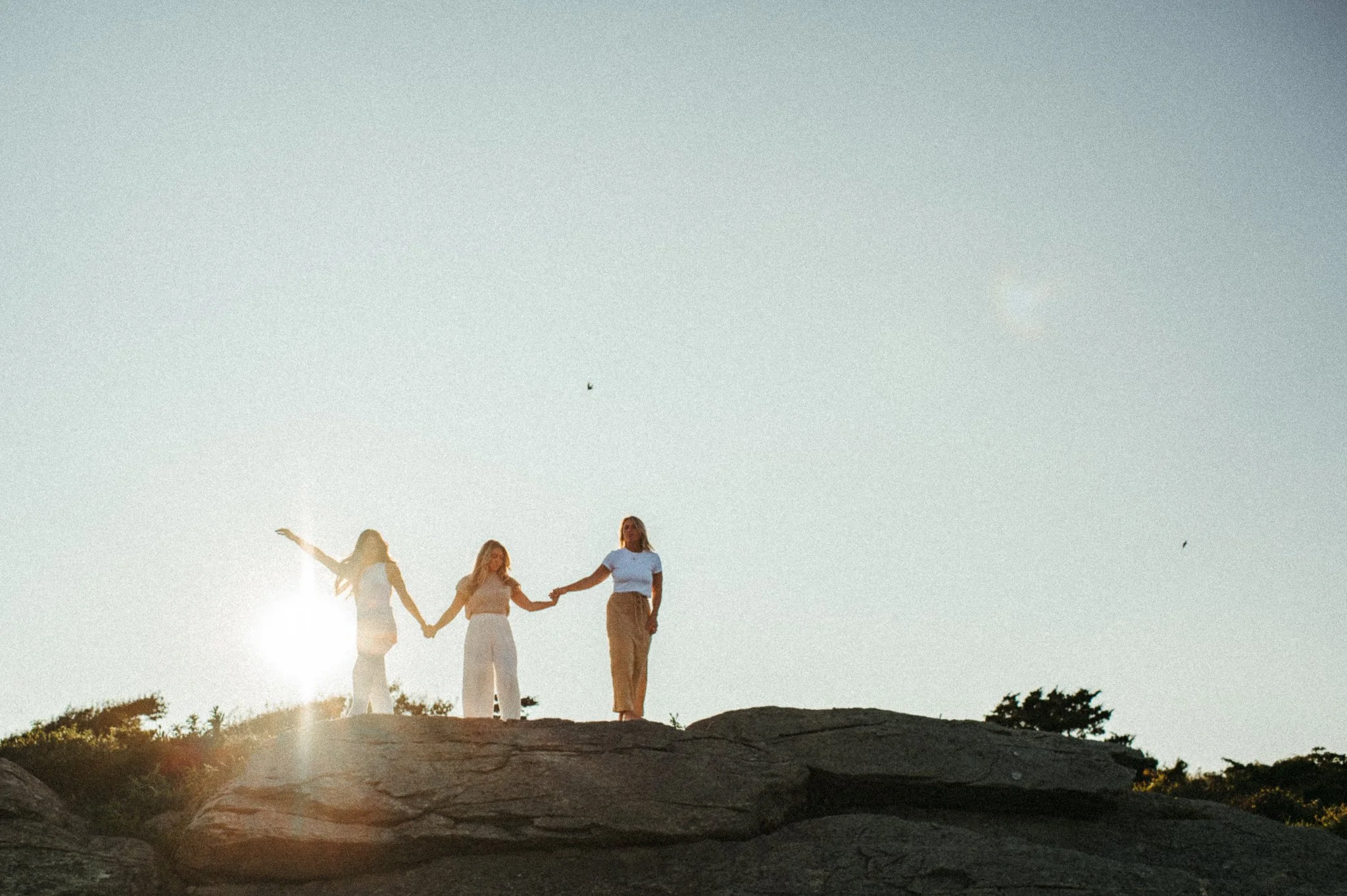 Three women holding hands on a rocky hill at sunset or sunrise, with the sun visible behind them.