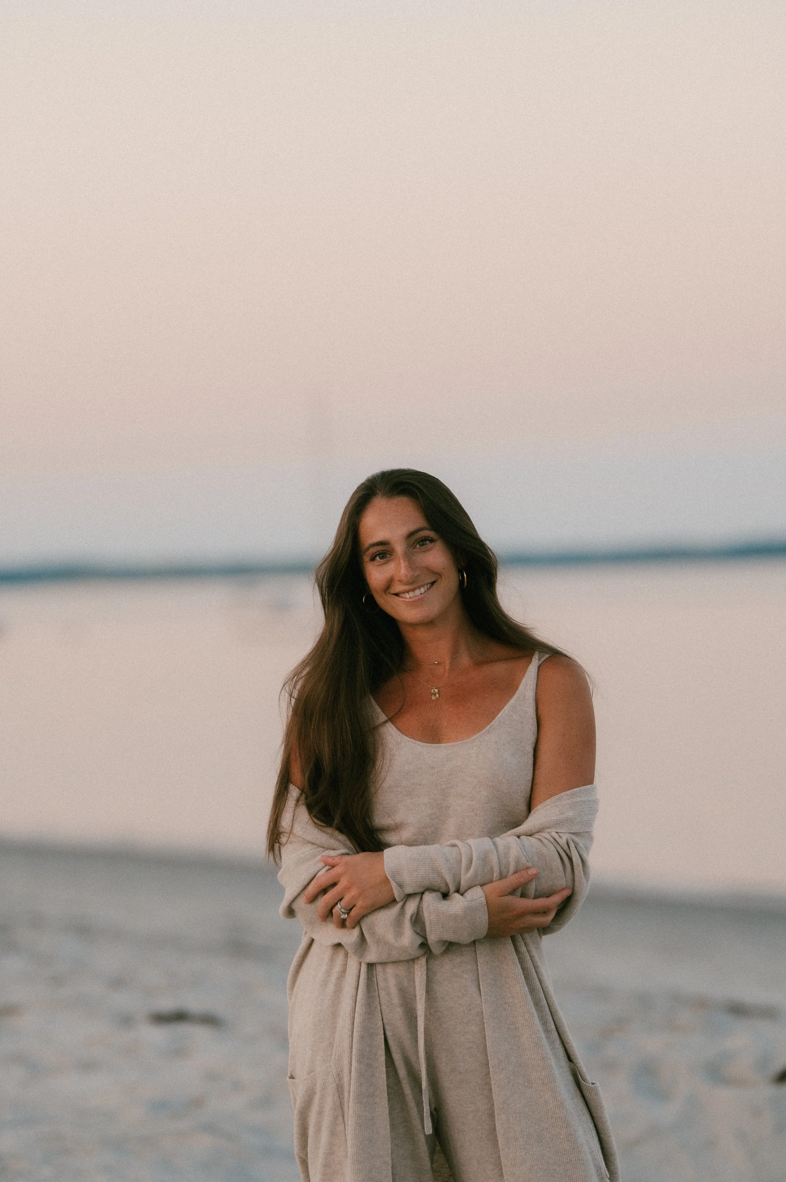 A woman smiling on a beach during sunset, wearing a tank top and a long cardigan.