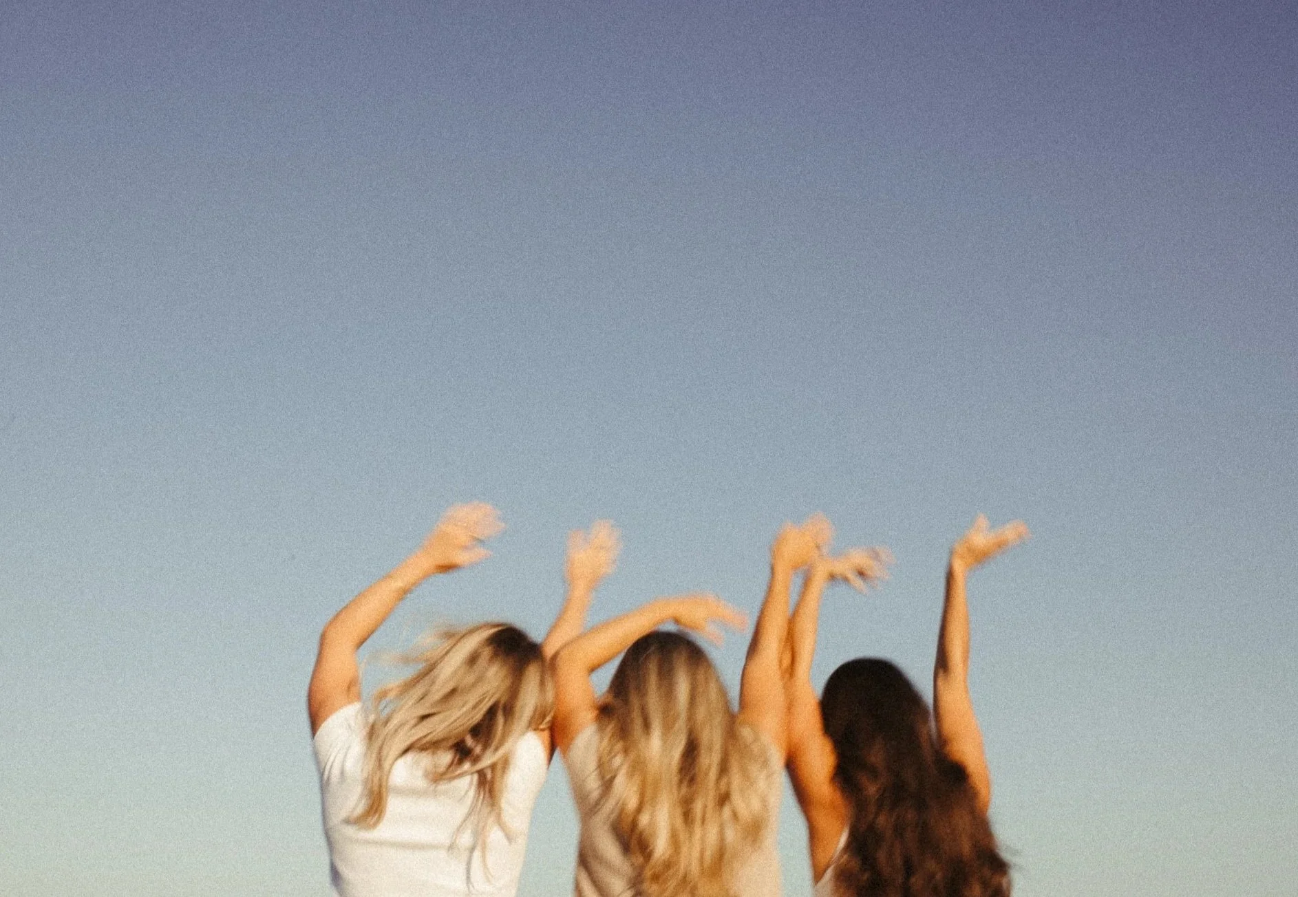 Three women with long hair, seen from behind, with their arms raised and hair flowing, standing outdoors under a clear sky.