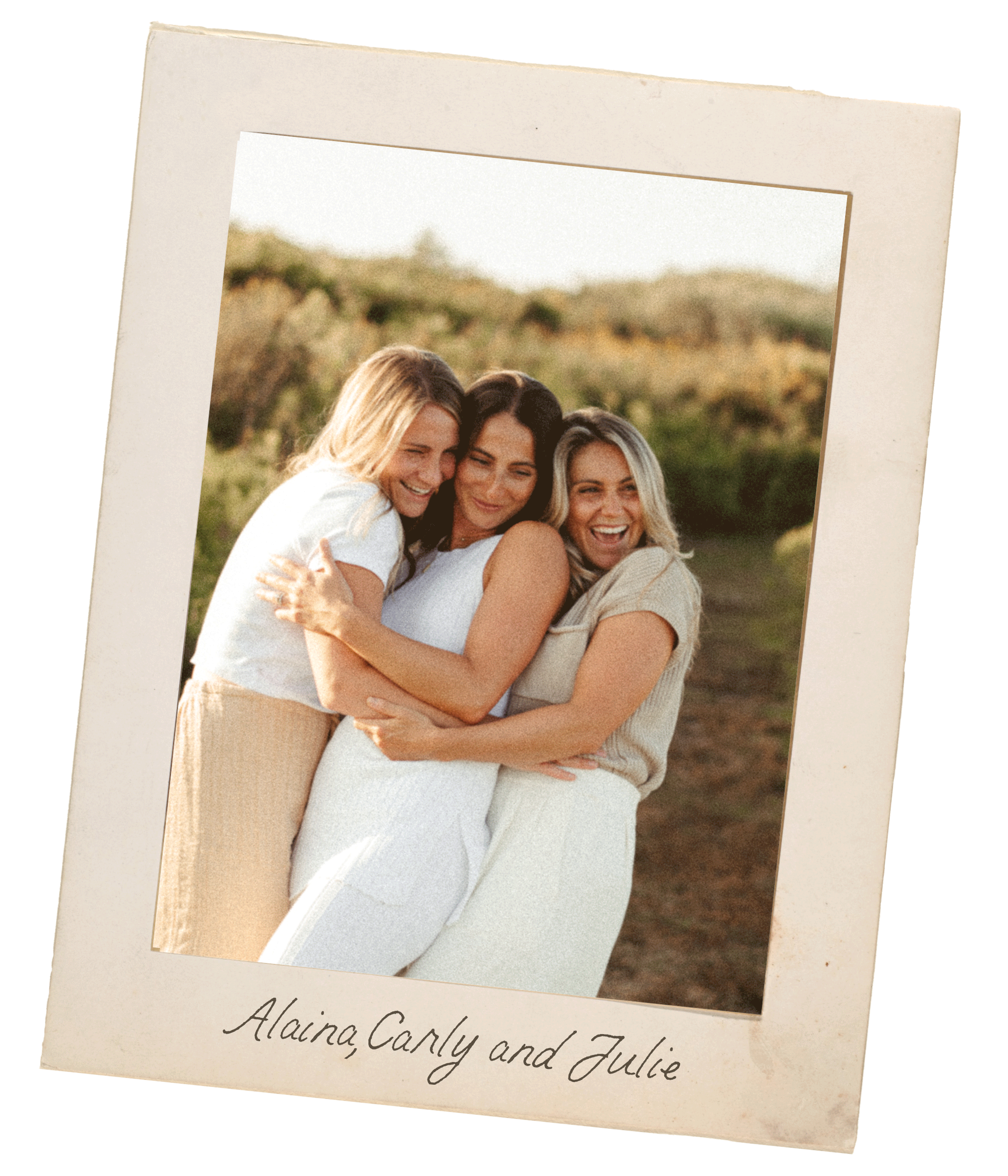 Three women hugging and smiling outdoors on a sunny day, with trees and grass in the background. The photo has a vintage look with a white border and handwritten text at the bottom reading "Alaina, Carly and Julie."