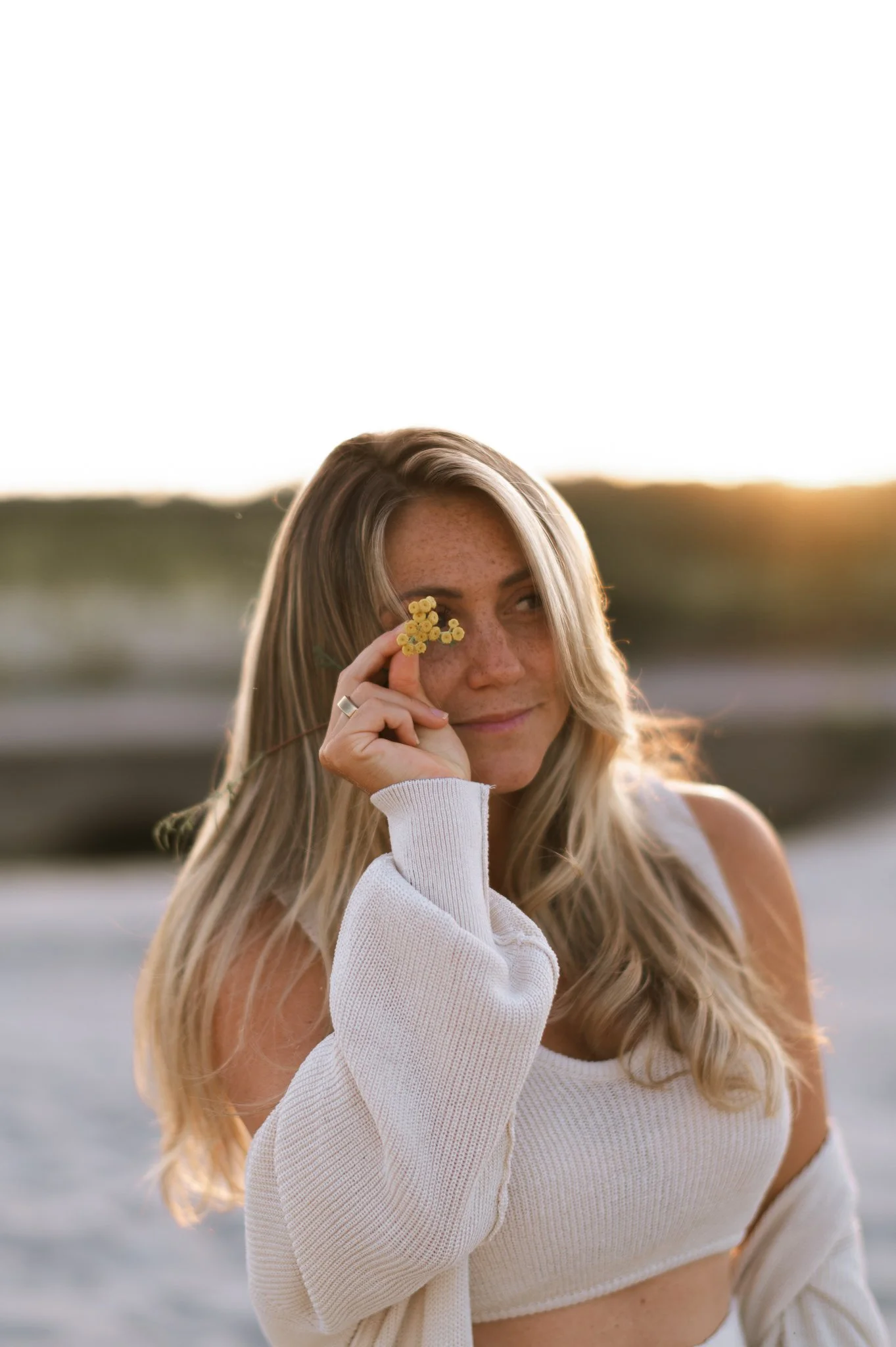 A woman with long blonde hair holding a small yellow flower near her eye outdoors, with a blurred natural background and soft sunlight.