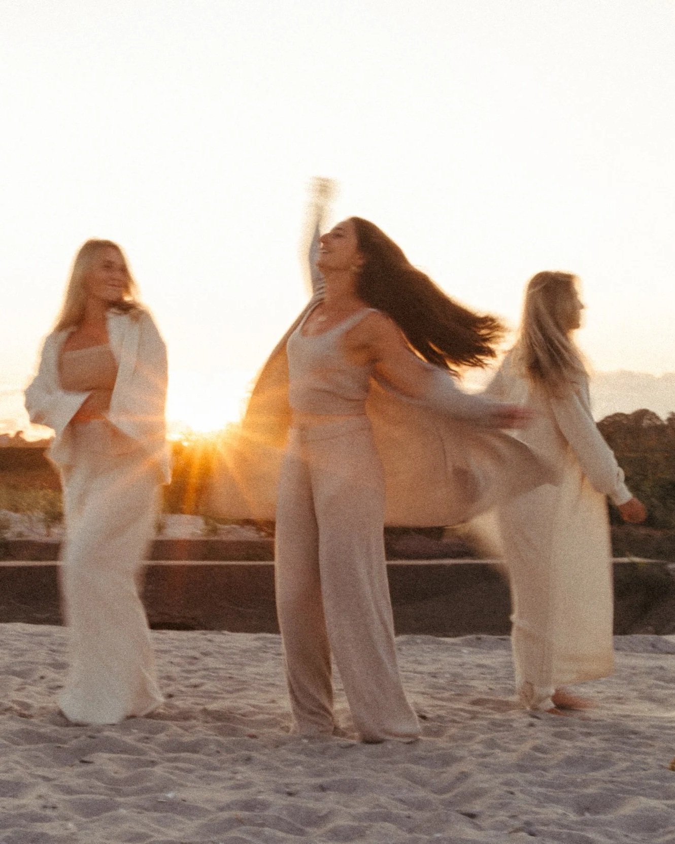 Three women dancing on the beach at sunset, wearing light-colored clothing.