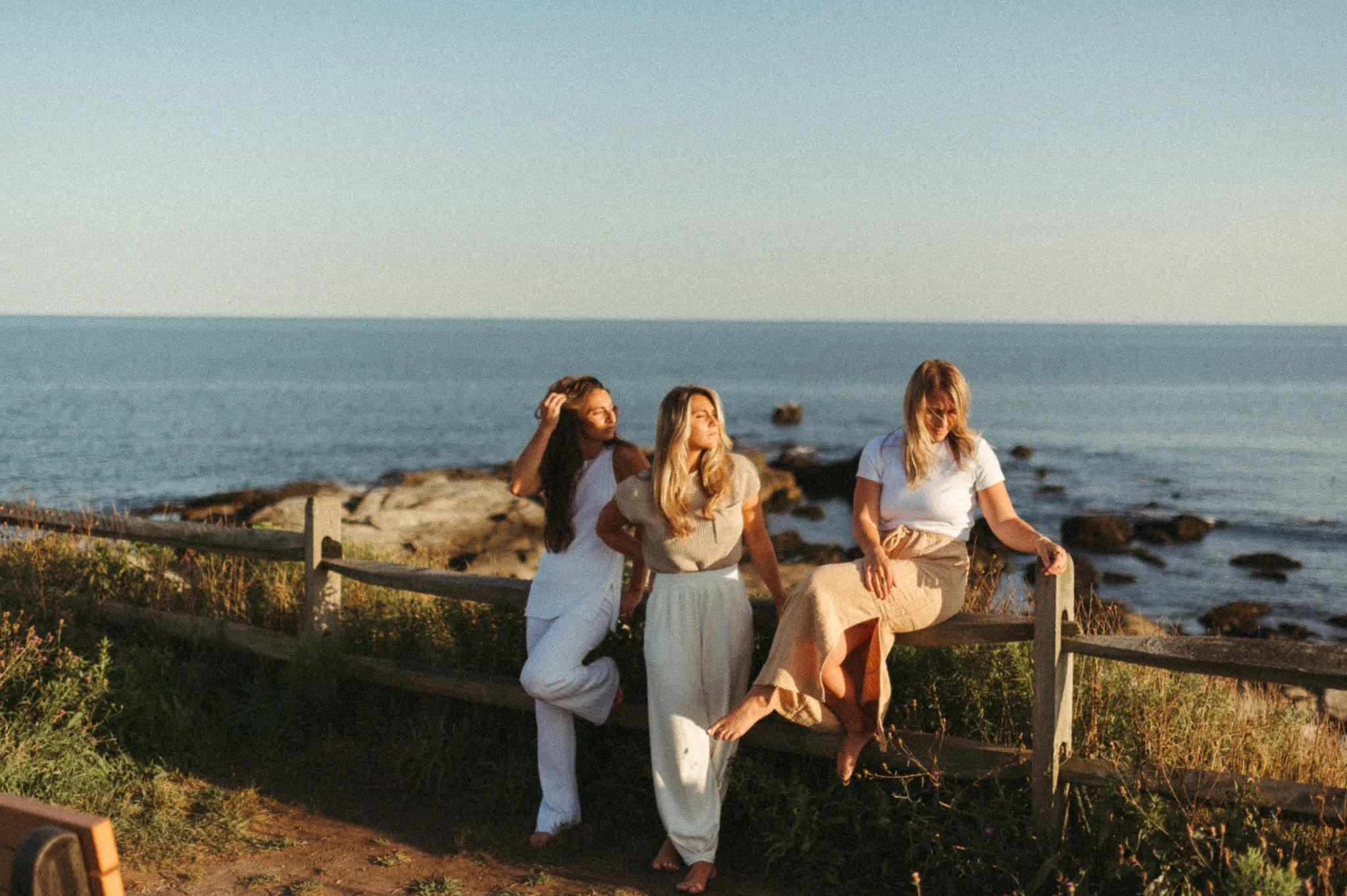 Three women with blonde and brunette hair at the seaside, sitting and standing on a wooden fence, dressed in light-colored casual clothing, during sunset or late afternoon.