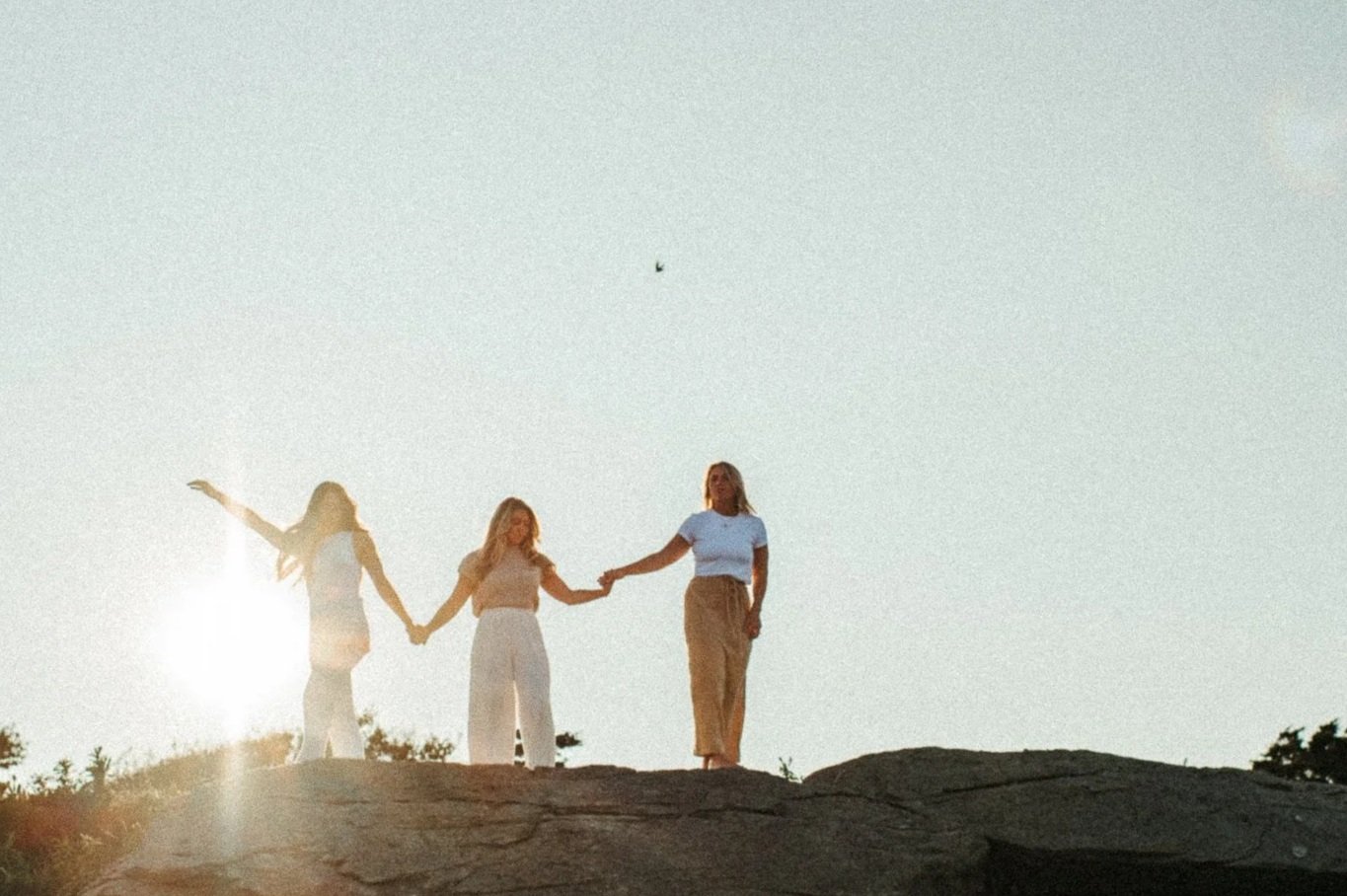 Three women holding hands on a rocky hill during sunset, with the sun in the background and a bird flying overhead.