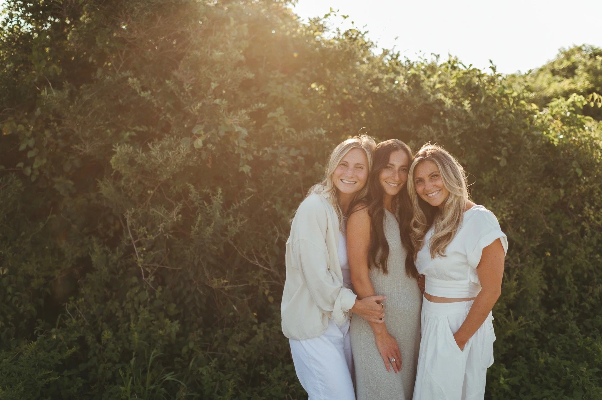Three women smiling outdoors in casual white and light-colored clothing, standing close together with greenery and sunlight in the background.