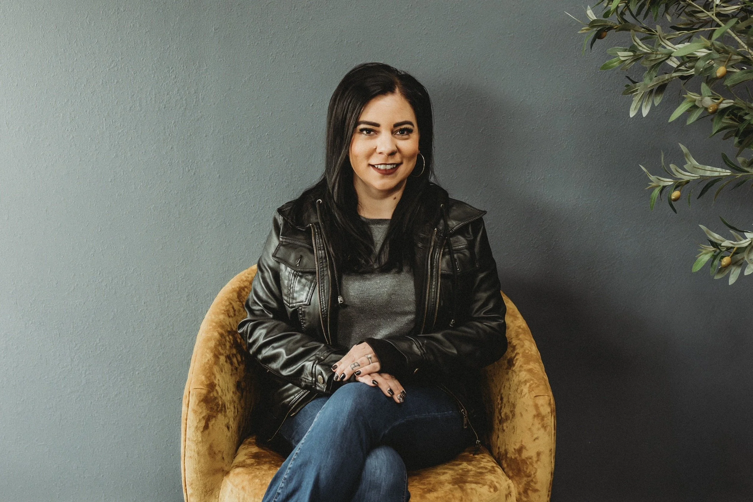 A woman with dark hair, wearing a black leather jacket, sitting on a mustard-colored velvet armchair, smiling, with a dark gray wall and a plant in the background.