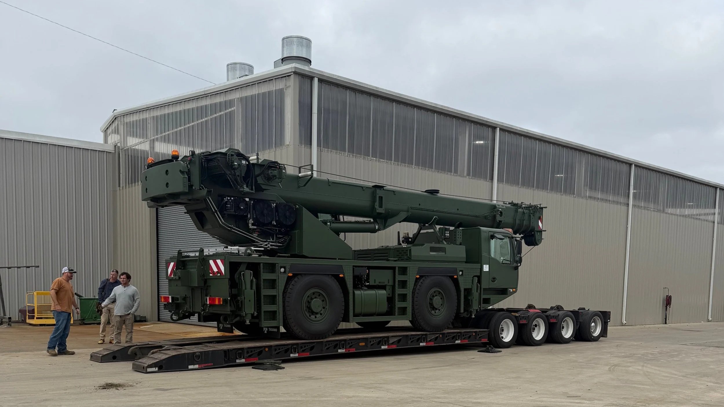 A large green military crane truck on a flatbed trailer outside a metal industrial building, with three men standing nearby.