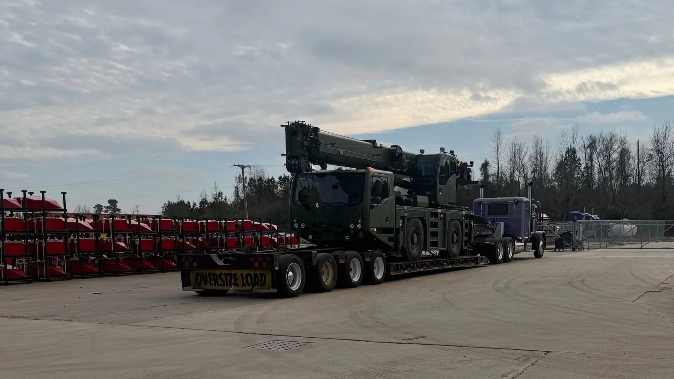 A large flatbed truck carrying a military crane is parked outside. In the background, red and black traffic light structures are stacked, with some trees and utility poles visible under a cloudy sky.