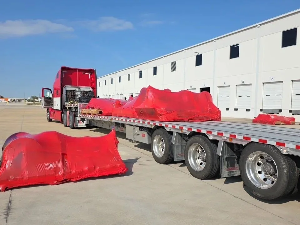 A red semi-truck with a flatbed trailer parked in an industrial area. Large red objects wrapped in plastic are loaded on the trailer, with some placed on the ground nearby. A white warehouse building with multiple garage doors is visible in the backg