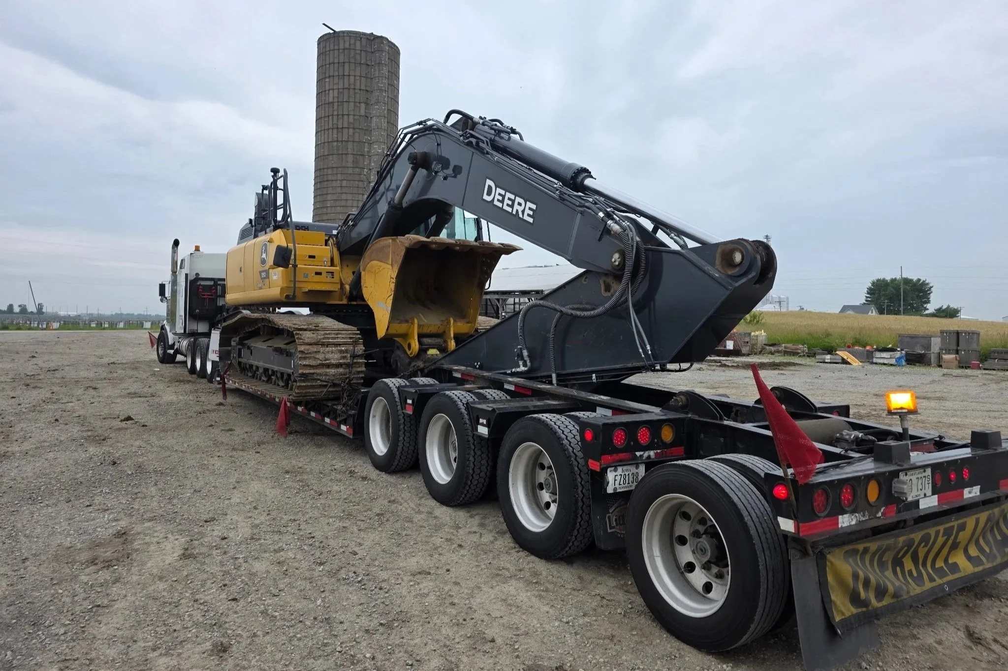 A large John Deere excavator loaded onto a flatbed truck, parked on a gravel lot with a grain elevator and rural landscape in the background.