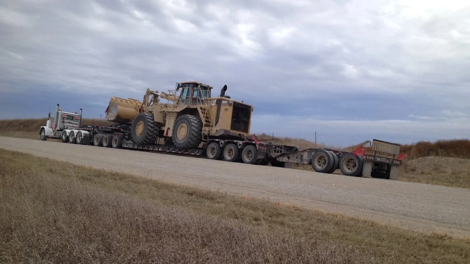 A large heavy machinery vehicle, a loader, on a flatbed truck being transported on a highway with grassy fields and a cloudy sky in the background.