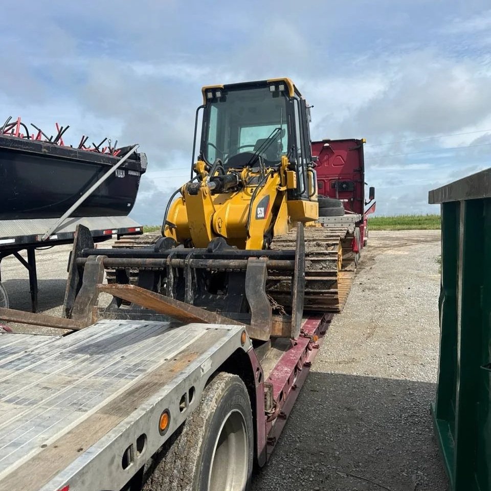 A yellow compact excavator loaded on a flatbed trailer with other equipment around on a gravel lot under a cloudy sky.