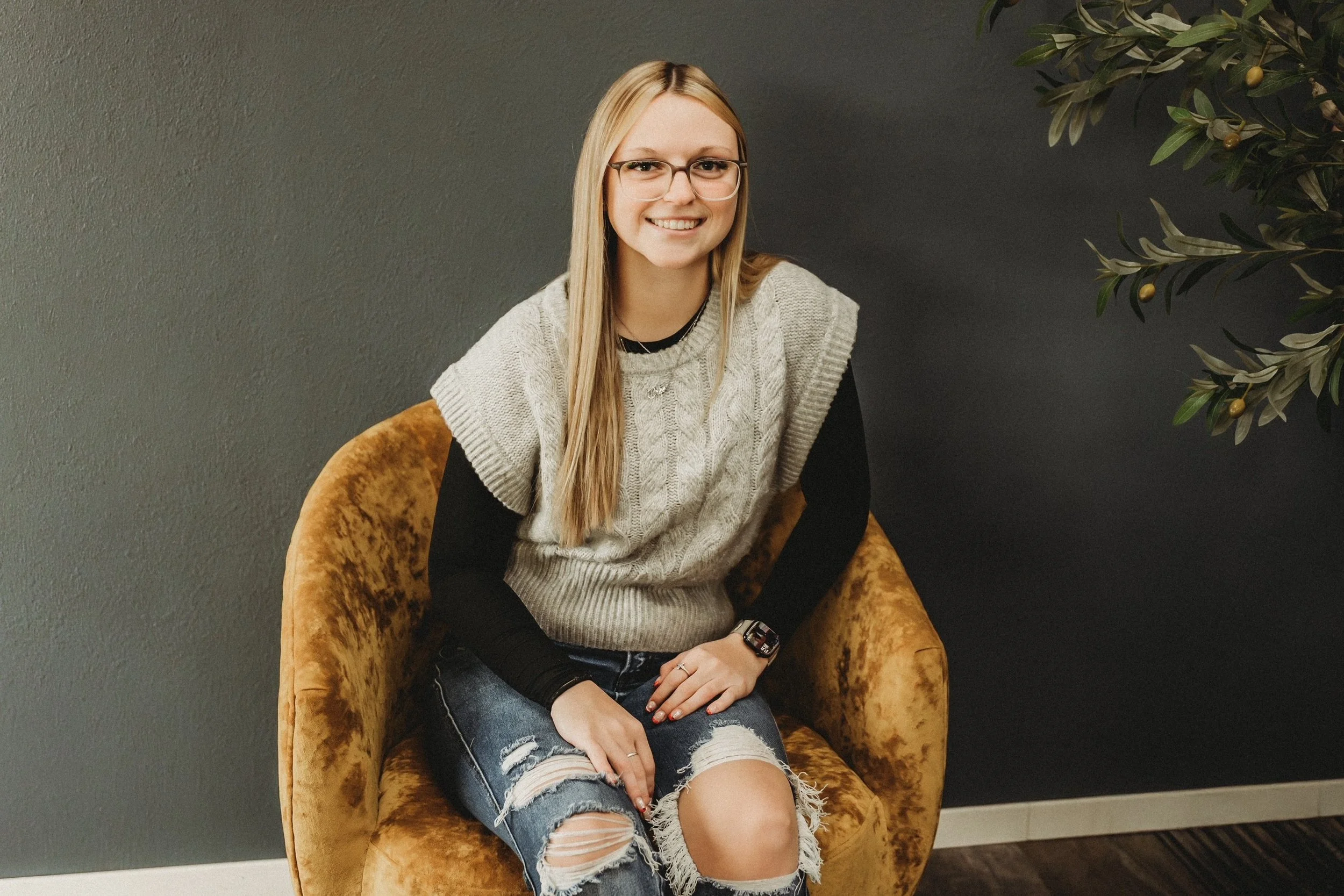 A young woman with long blonde hair, glasses, and a big smile sitting on a brown velvet armchair next to a green potted plant, wearing a gray knit vest over a black long sleeve shirt and ripped jeans.