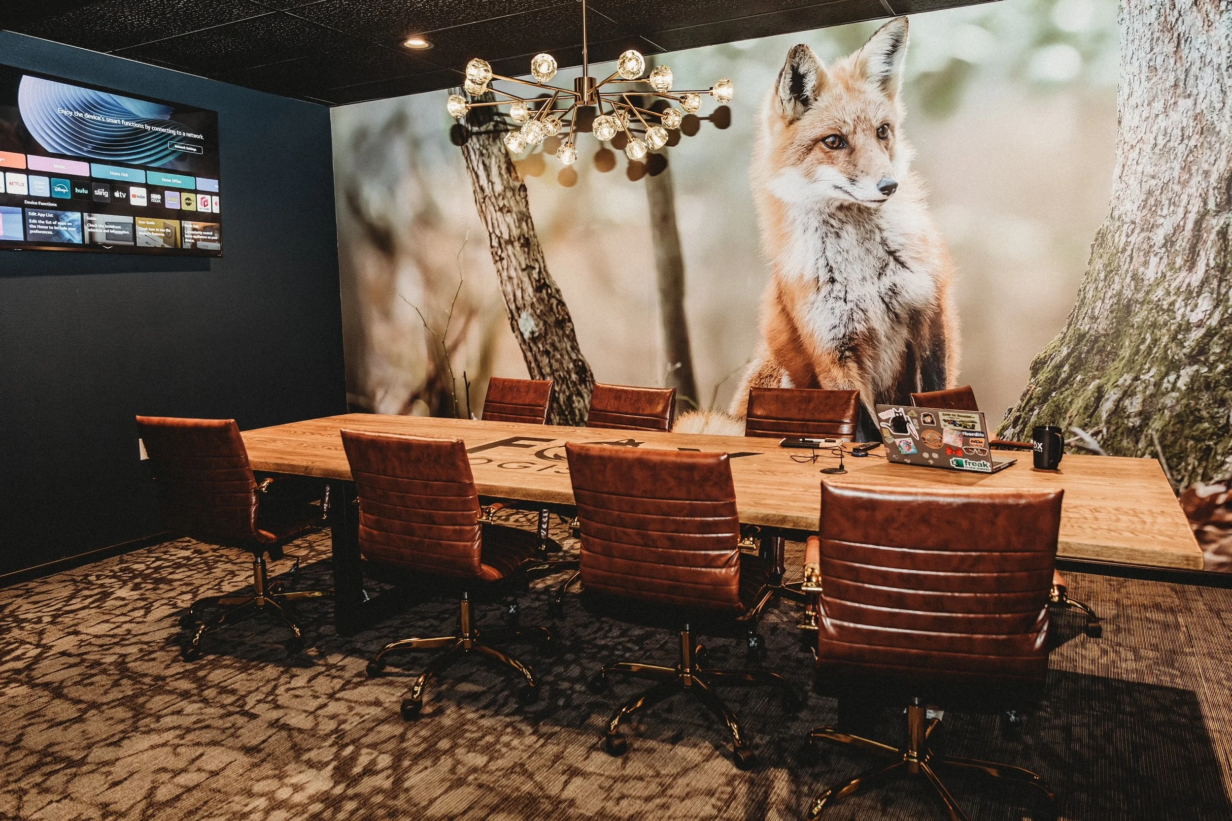 A conference room with a large wooden table surrounded by six brown leather chairs. The wall has a large mural of a fox sitting next to a tree in a forest setting. A modern chandelier with multiple bulbs hangs from the ceiling, and a television screen displaying streaming service icons is mounted on a black wall.