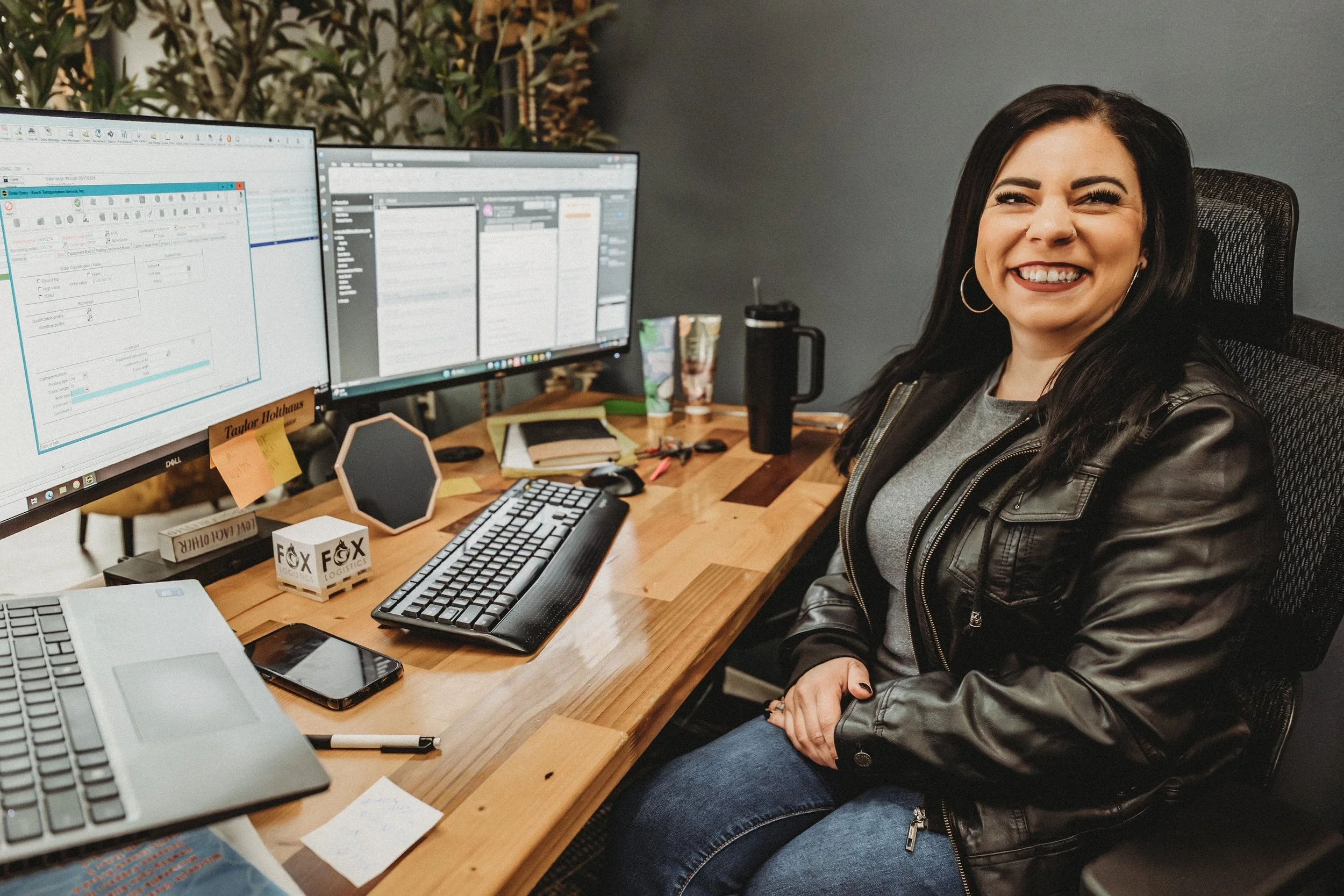 A woman sitting at a desk with two computer monitors, smiling at the camera, with office supplies and personal items on her desk.