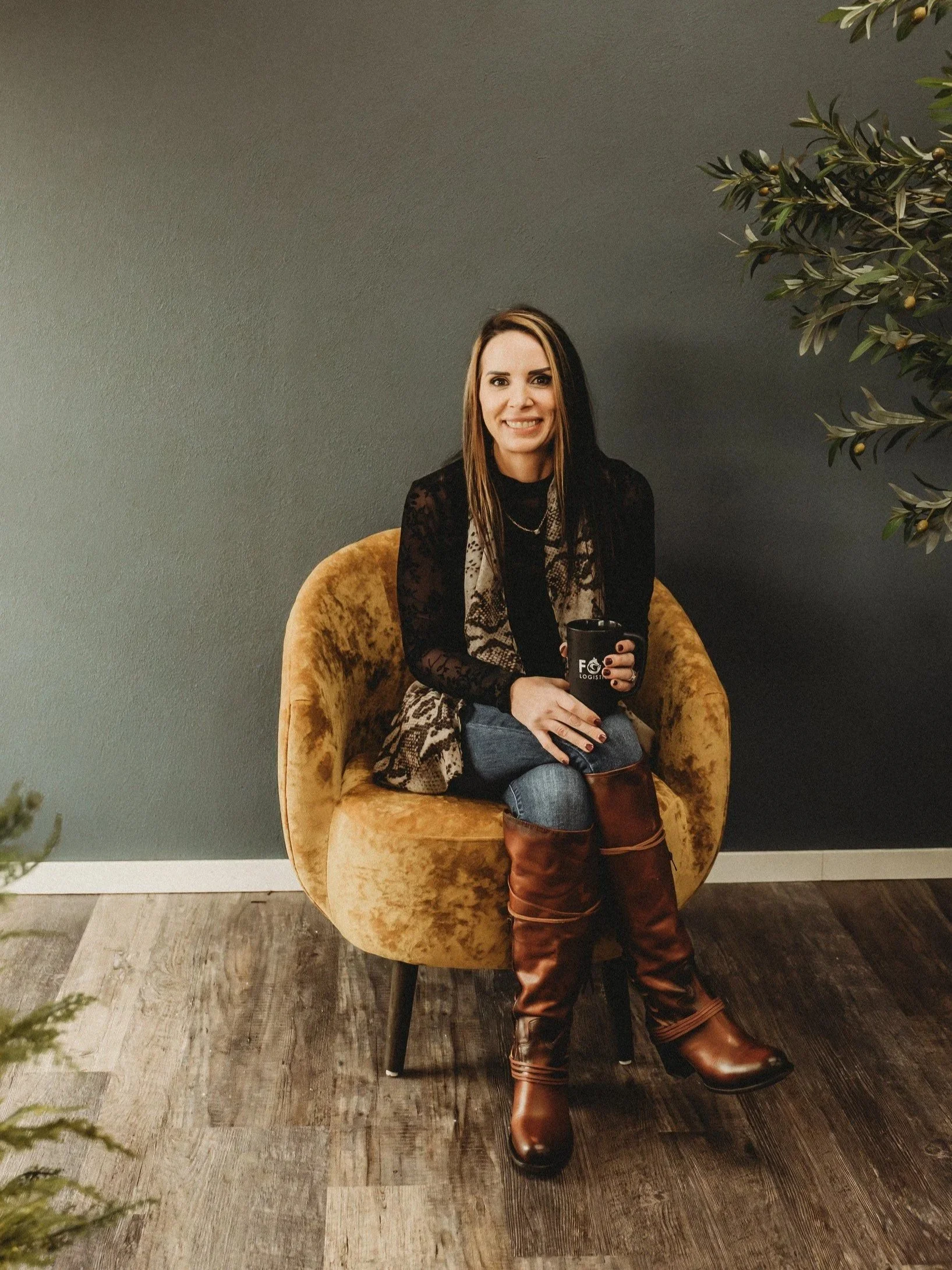 A woman sitting in a mustard-colored velvet chair, holding a black mug, smiling at the camera.